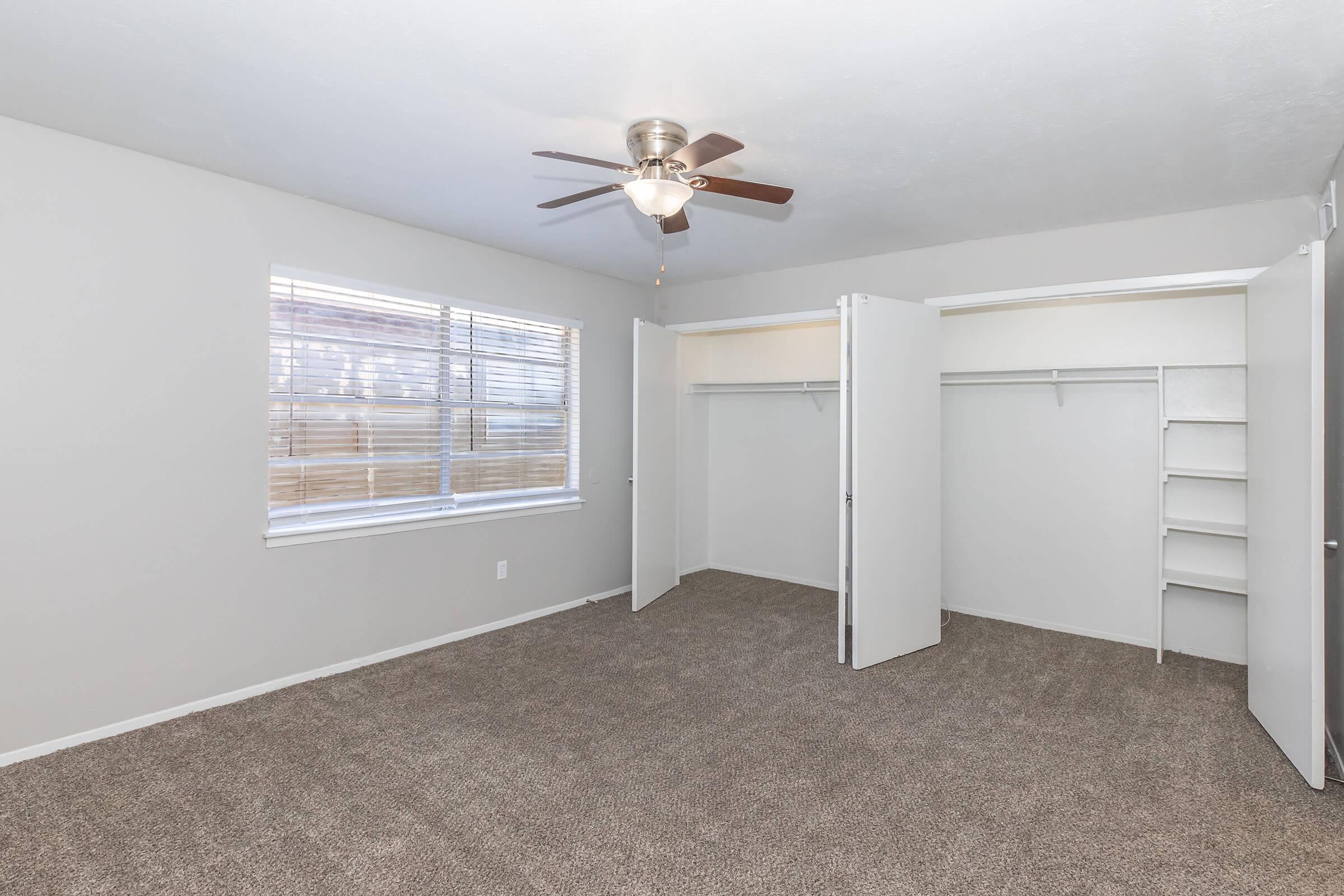 A spacious, empty bedroom featuring light gray walls, a ceiling fan, and large windows with blinds. The room has a plush carpet and two open closets with shelving, providing ample storage space. Natural light brightens the area, creating a welcoming atmosphere.