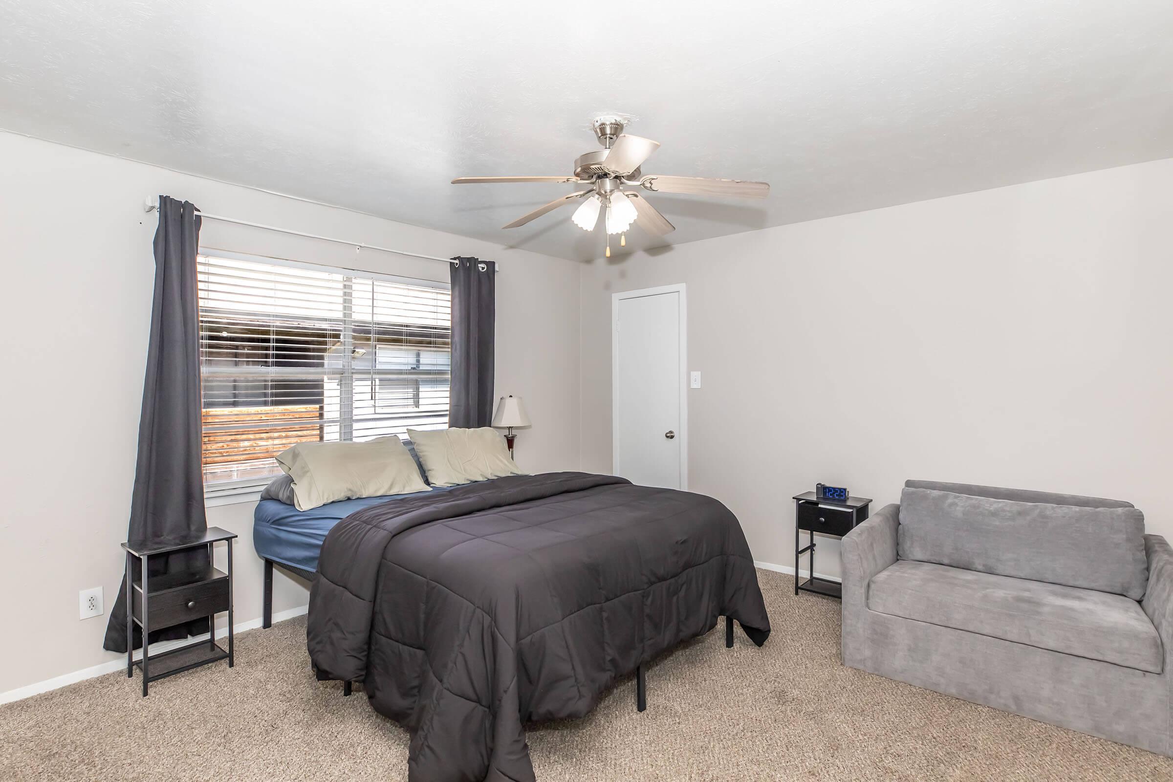A cozy bedroom featuring a queen-sized bed with dark bedding, a gray couch, and a small nightstand with a lamp. The room has light-colored walls and a window with curtains, allowing natural light to enter. A ceiling fan is also present, enhancing the comfortable atmosphere.