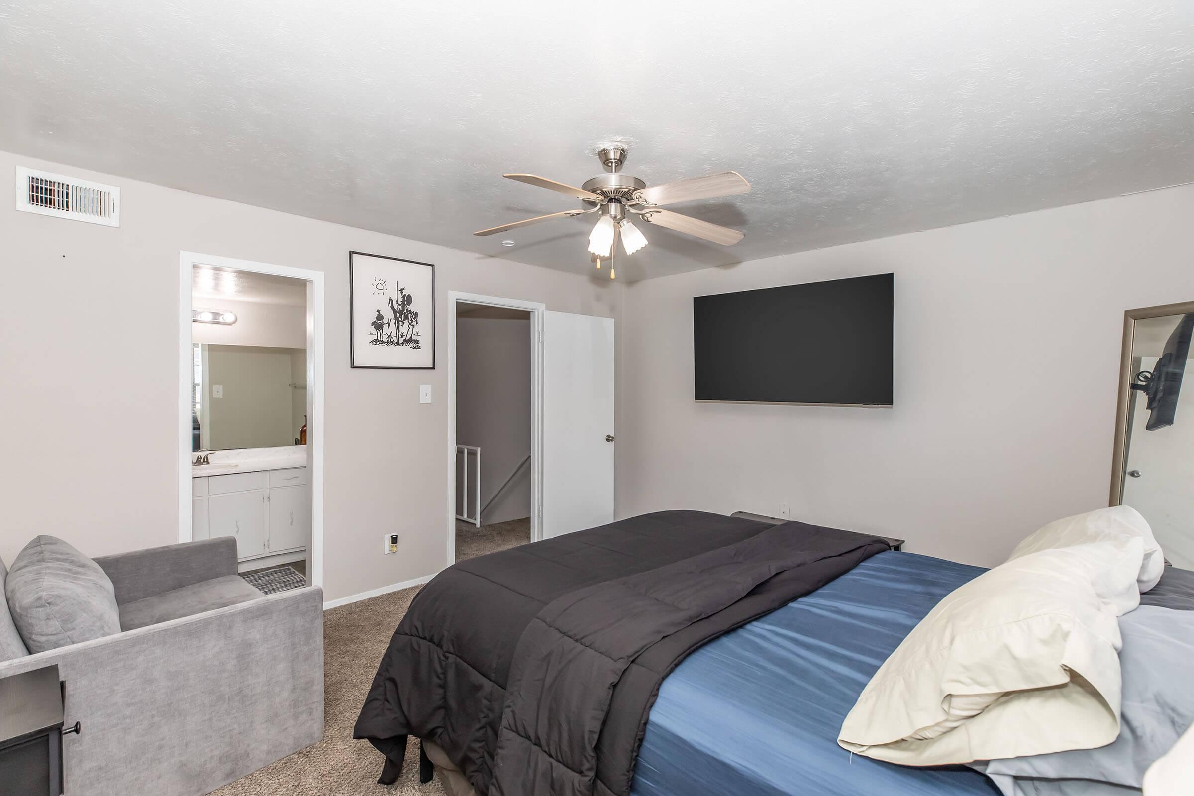 A cozy bedroom featuring a ceiling fan, a large flat-screen TV mounted on the wall, a bed with a dark comforter and light-colored pillows, and a small gray armchair. There is a mirror and a door leading to a bathroom area in the background, with neutral-colored walls and carpet.