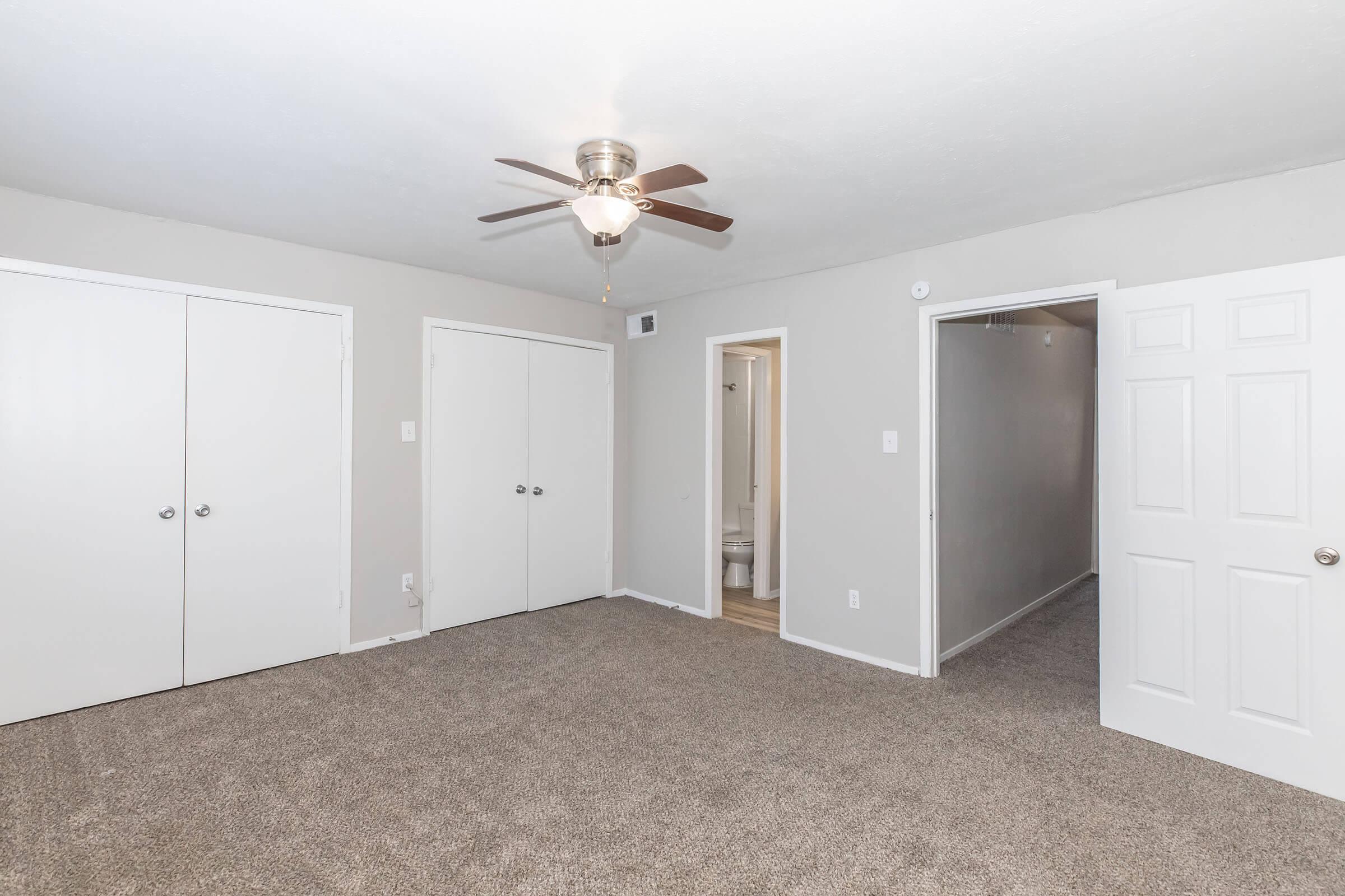 A spacious, well-lit room with light gray walls and a ceiling fan. The floor is covered in soft, beige carpet. Two closed white doors are visible on the left, while another door leads to a bathroom on the right. The overall atmosphere is clean and inviting.