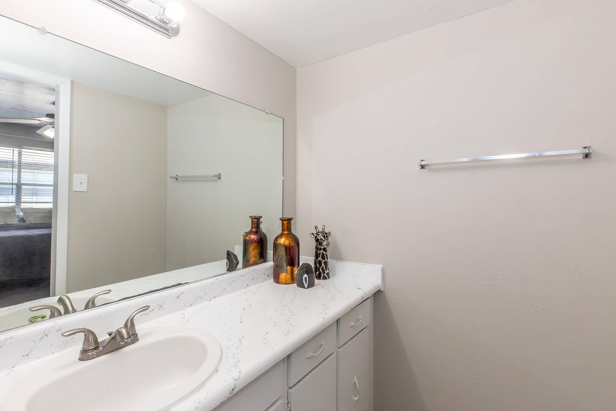 A clean bathroom with a white countertop and sink, featuring two decorative brown vases and a small figurine on the countertop. A large mirror reflects the space, and a towel rack is mounted on the wall. Natural light enters from a nearby window, enhancing the neutral color scheme.