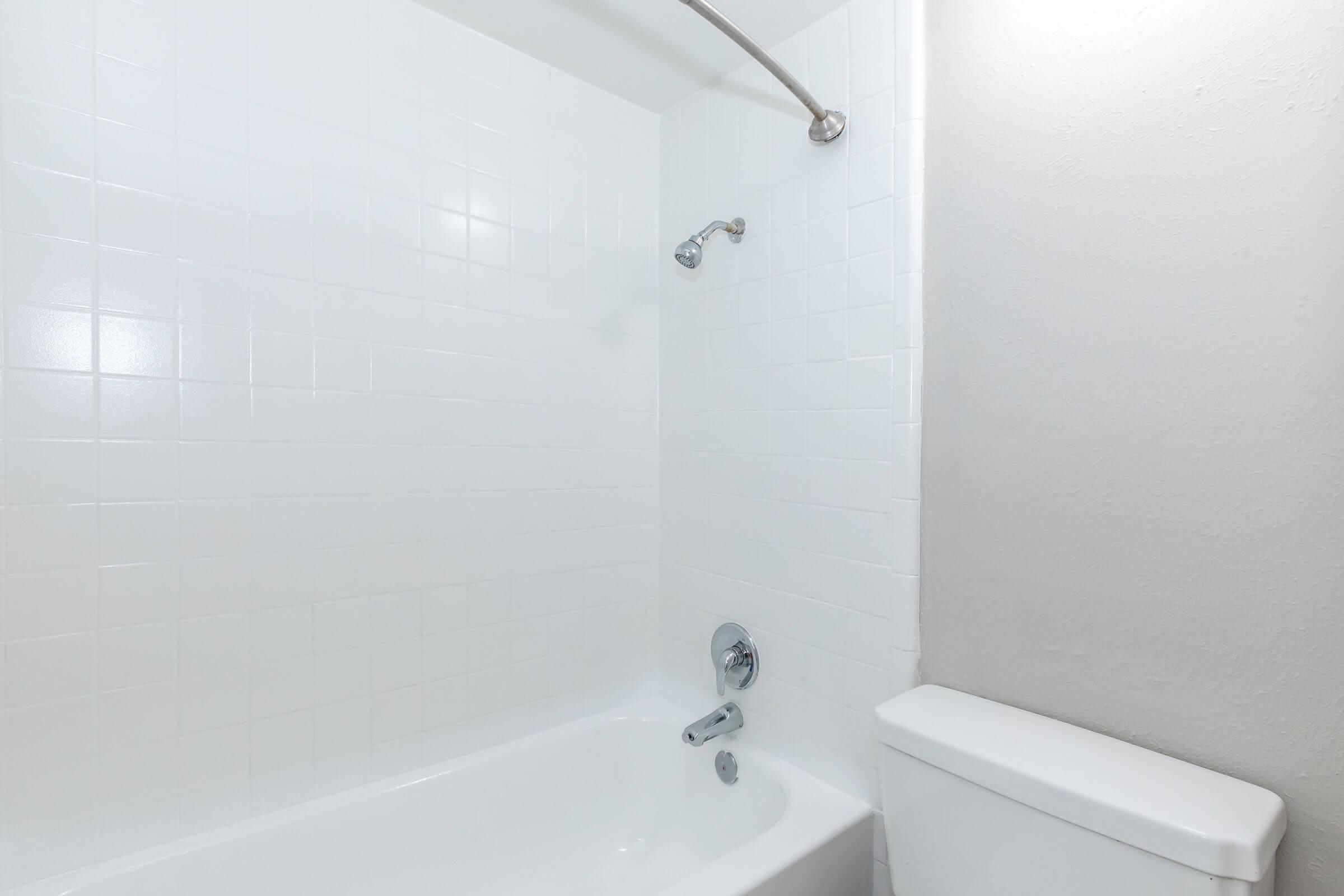 A clean, modern bathroom featuring a white bathtub with a showerhead and a single faucet. The walls are tiled in white, and there's a toilet next to the bathtub. The overall design is simple and minimalistic, creating a bright and airy atmosphere.