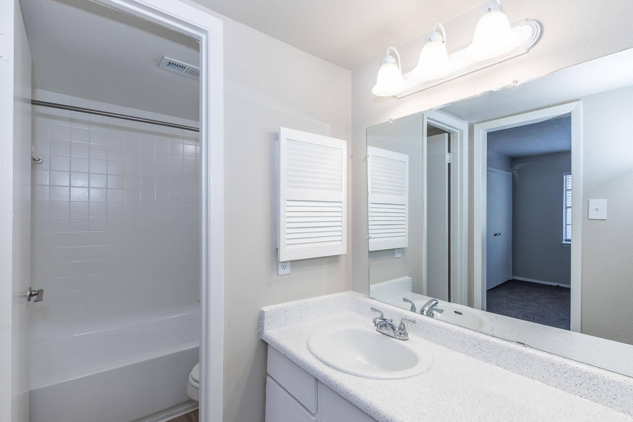 A clean, modern bathroom featuring a white bathtub and shower combination, a light-colored countertop with a sink, a large mirror, and bright lighting fixtures. The walls are painted in a neutral tone, and a door leads to another room in the background, creating an open and spacious feel.