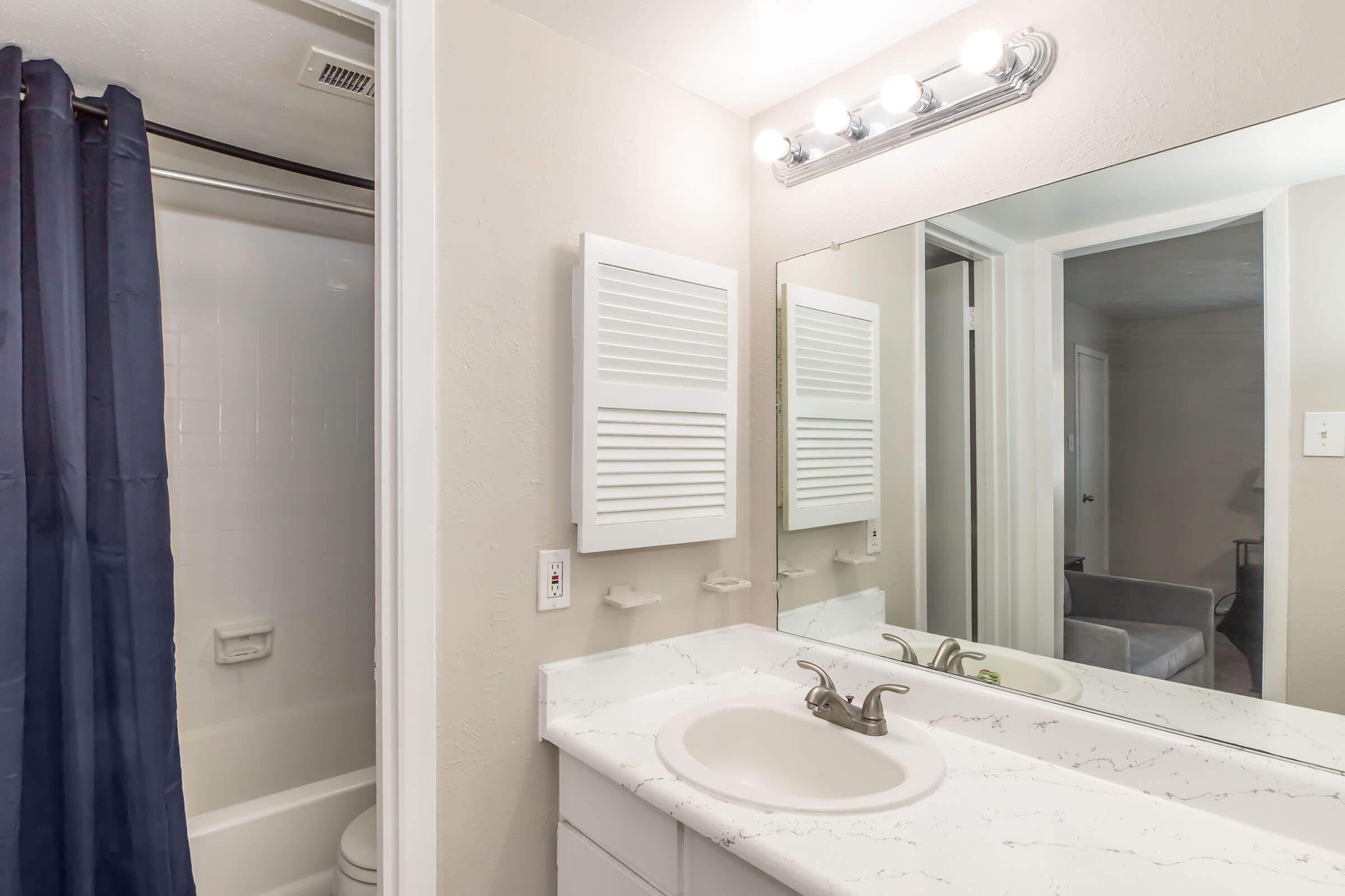 A well-lit bathroom featuring a white marble countertop with a sink, a large mirror, and a bathtub with a shower curtain. The walls are painted a light color, and there is a ventilated wall panel. Soft lighting from the overhead fixture adds to the ambiance.