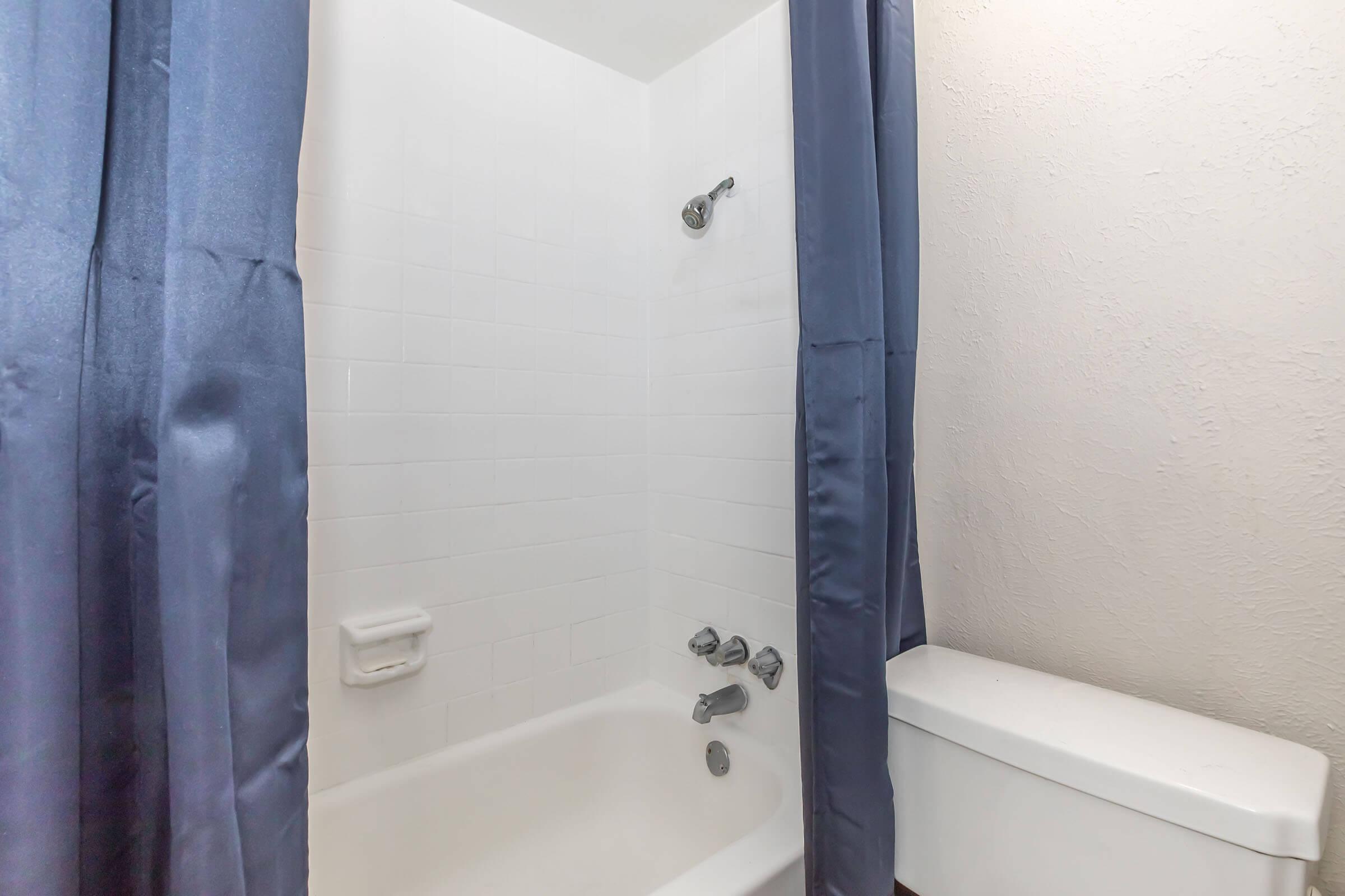 A bathroom featuring a shower with white tiles and a blue shower curtain. The tub area includes a showerhead and water controls. To the side, there is a white toilet against the wall. The overall setting is simple and clean, ideal for a functional bathroom space.