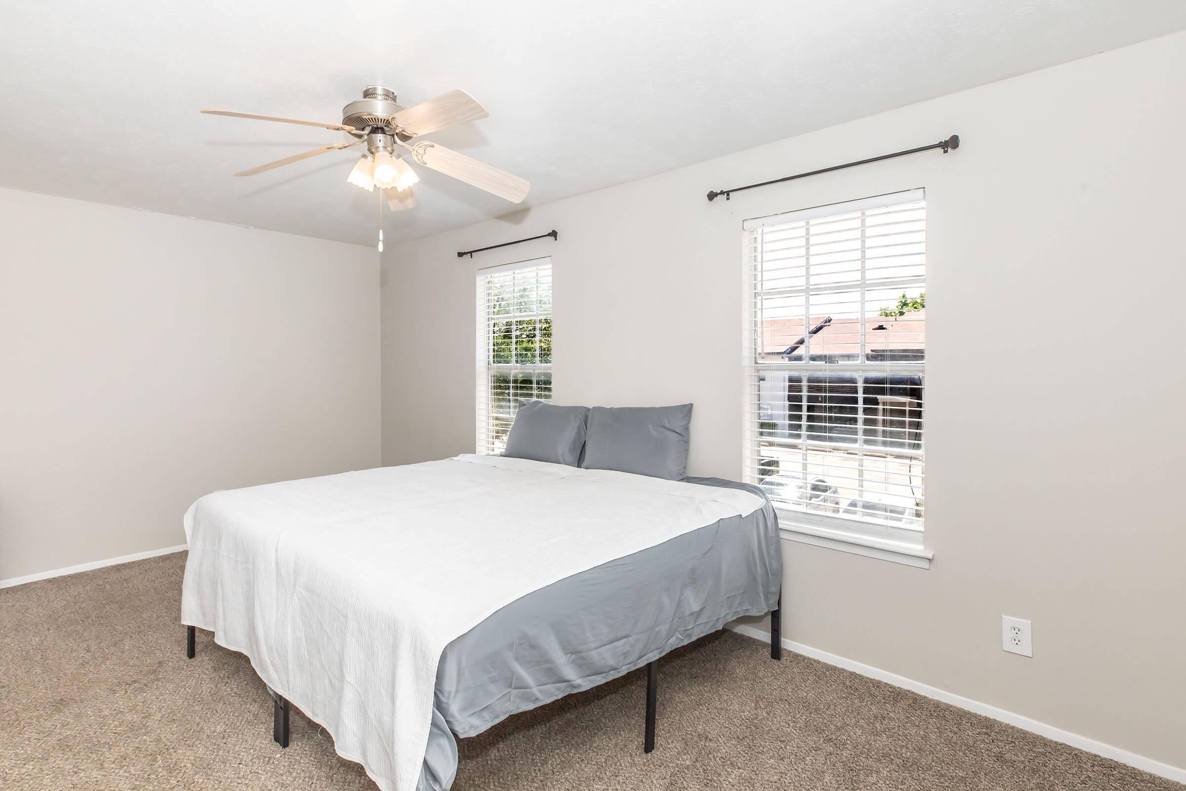 A cozy bedroom featuring a bed with gray bedding and two pillows, a ceiling fan with lights, and two windows with blinds providing natural light. The walls are painted in a light color, and the floor is carpeted, creating a warm and inviting atmosphere.