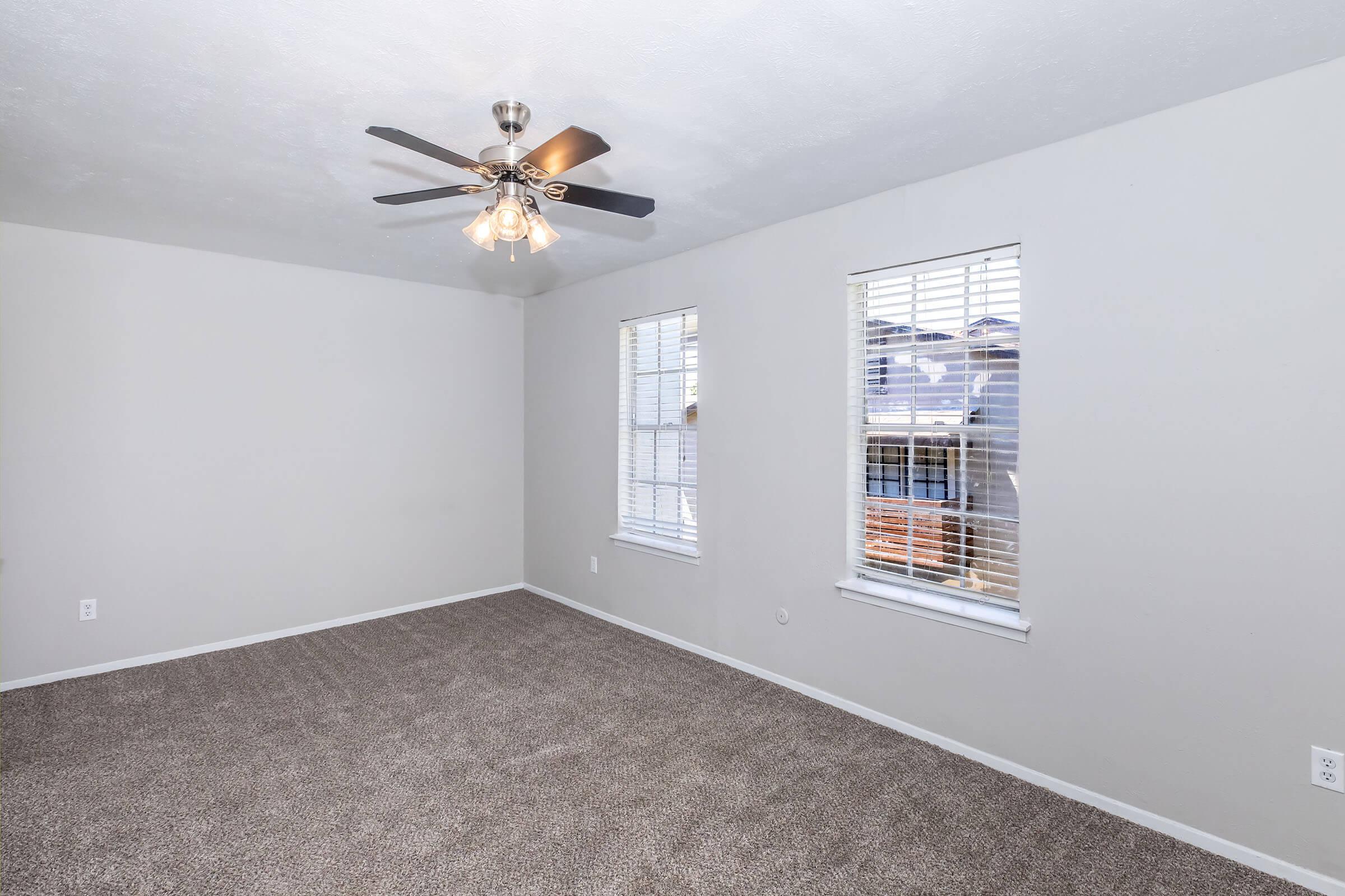 A well-lit room featuring light gray walls, a ceiling fan with five blades, and large windows with white blinds. The floor is covered with beige carpet, creating a cozy atmosphere. The room is empty, ideal for various furniture arrangements.