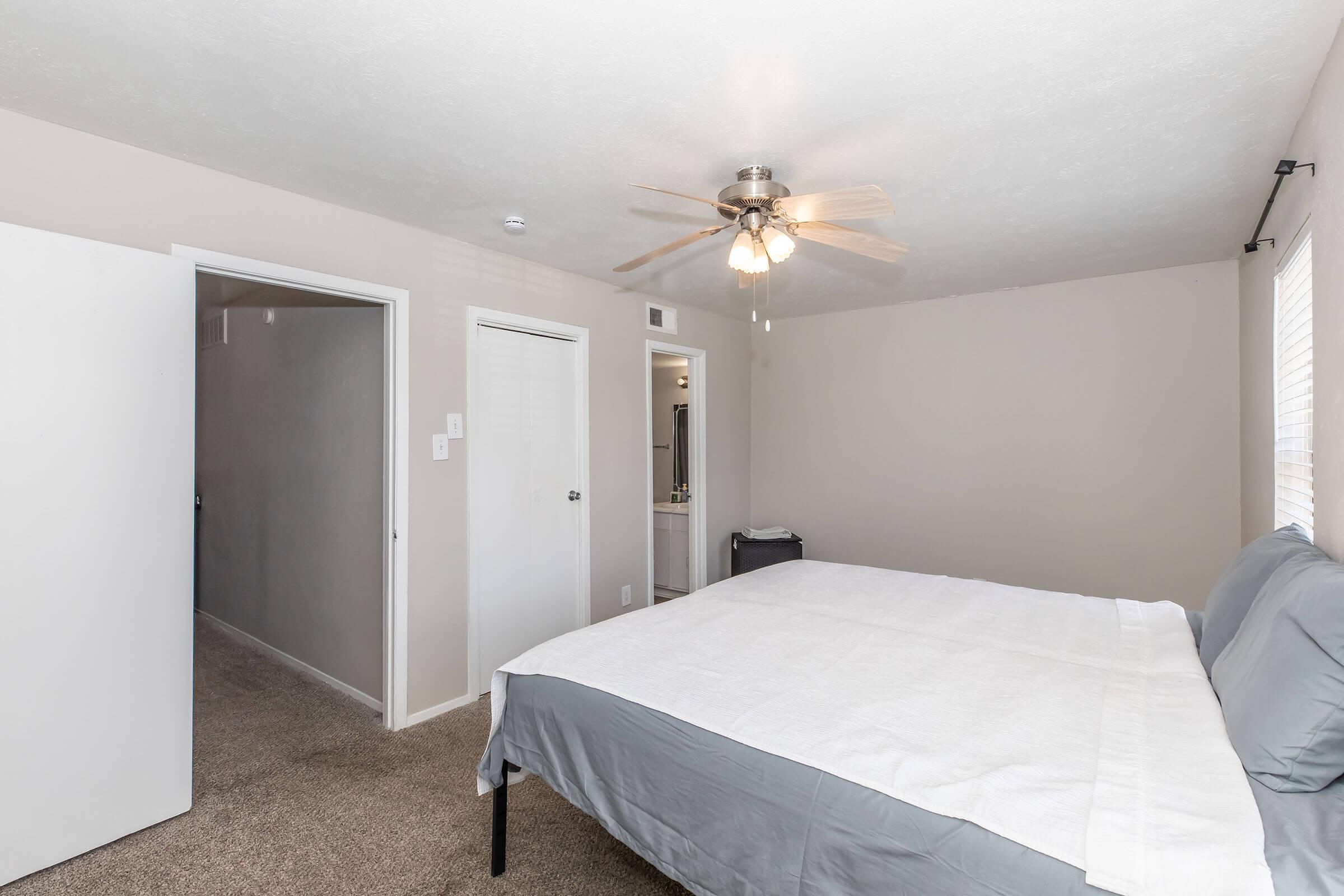 A well-lit bedroom featuring a bed with gray linens, a ceiling fan, light-colored walls, and carpeted flooring. The room has a doorway leading to a hallway and another door indicating a closet space. Natural light comes in through a window.