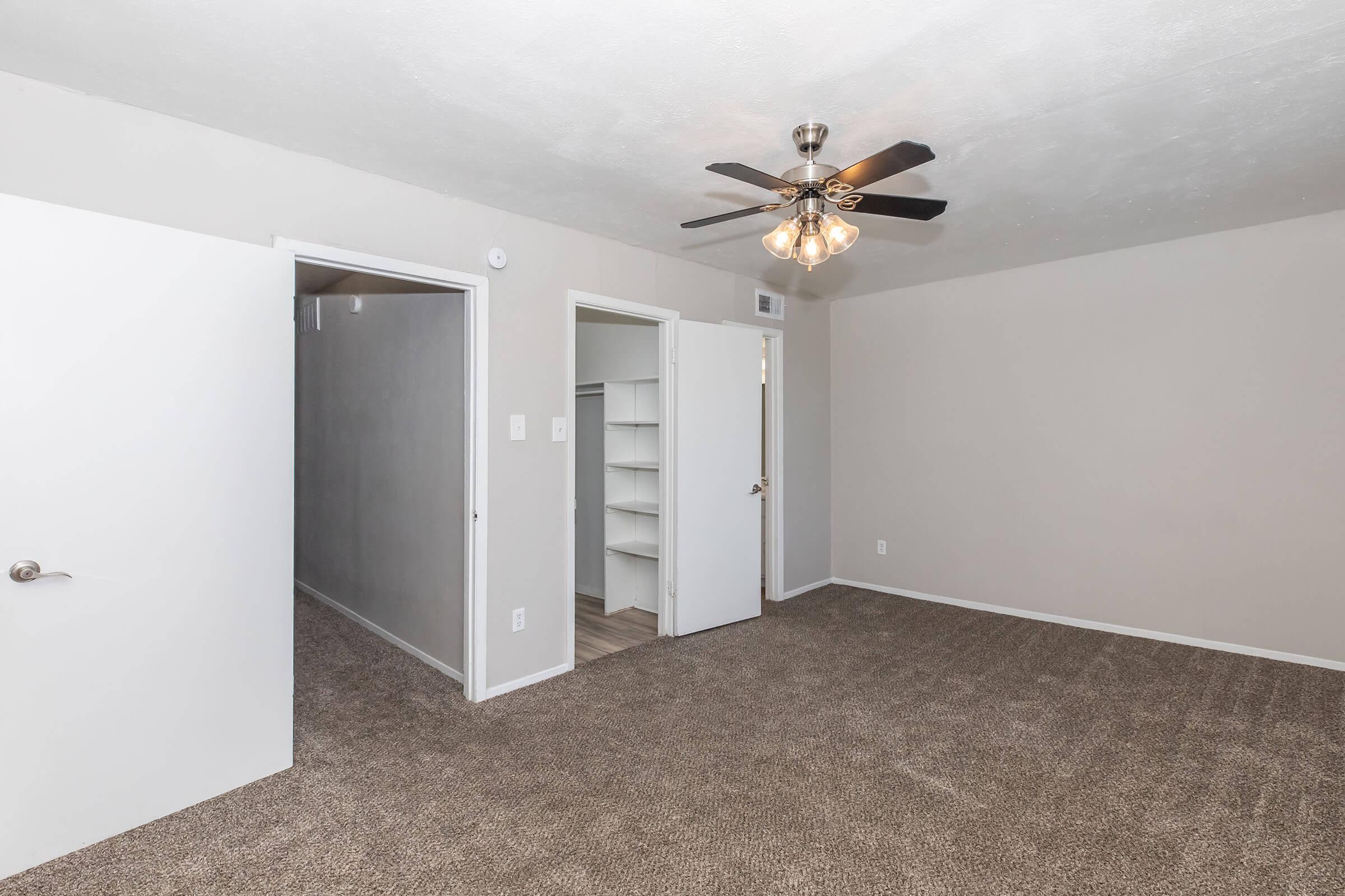 Interior view of a room featuring light brown carpet, a ceiling fan with lights, and two white doors leading to separate areas. One door opens to a closet or storage space, while the other leads to a hallway. The walls are painted in a neutral light color, creating a spacious and inviting atmosphere.