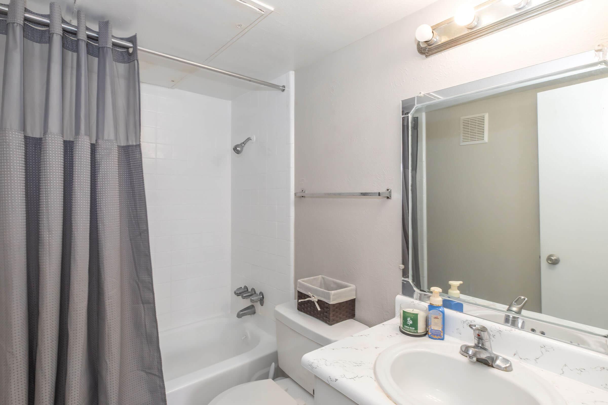 A small bathroom featuring a shower/tub combination with a gray curtain, a white sink with a marbled countertop, a mirror above the sink, and various toiletries on the counter. The walls are painted light gray, and a small basket is visible beside the toilet.