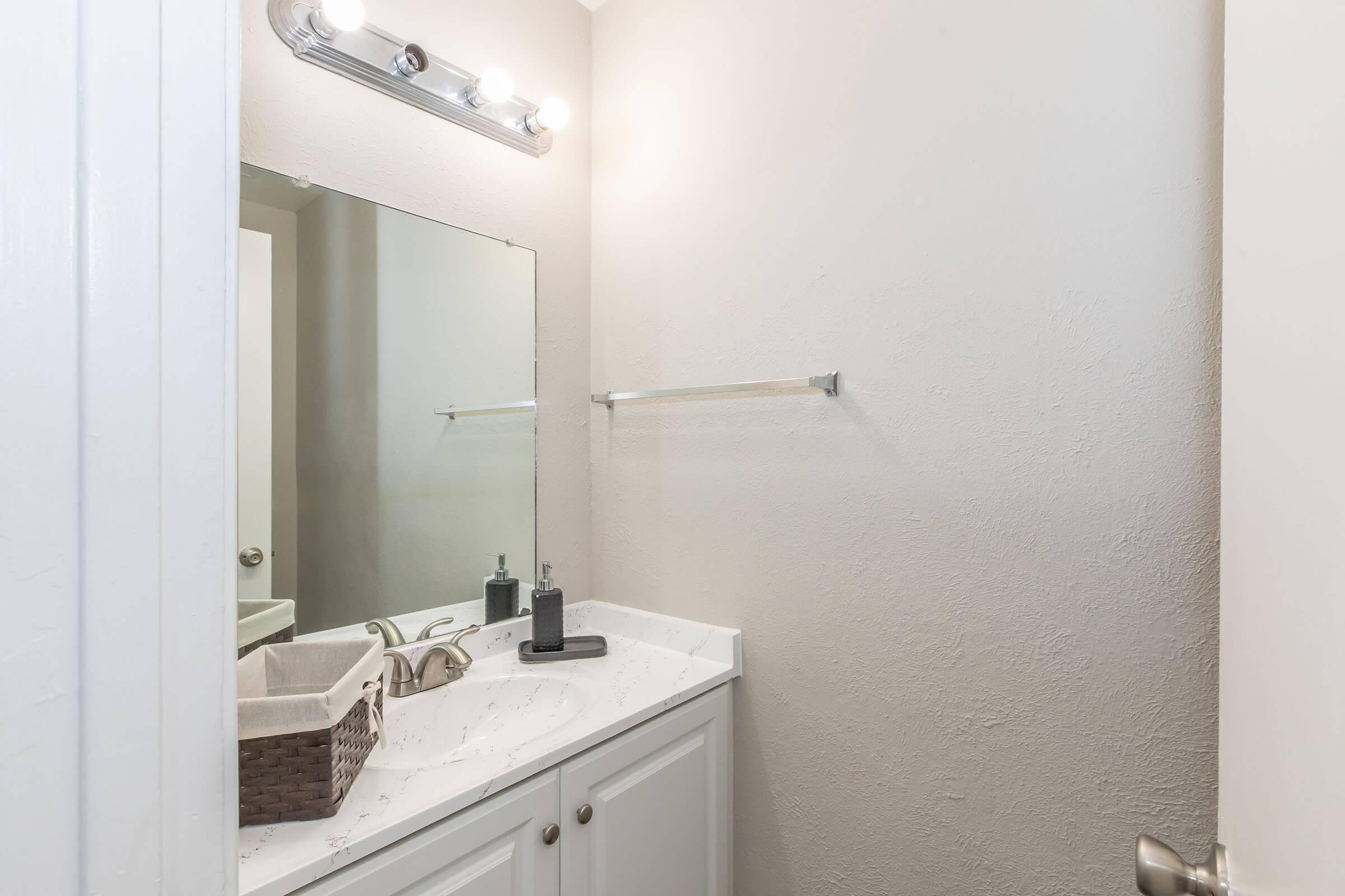 A clean, modern bathroom with a white cabinet below a marble countertop. A large mirror reflects the space, and there is a light fixture above. A towel bar is mounted on the wall, and a woven basket and a gray soap dispenser sit on the countertop, creating a minimalist aesthetic.