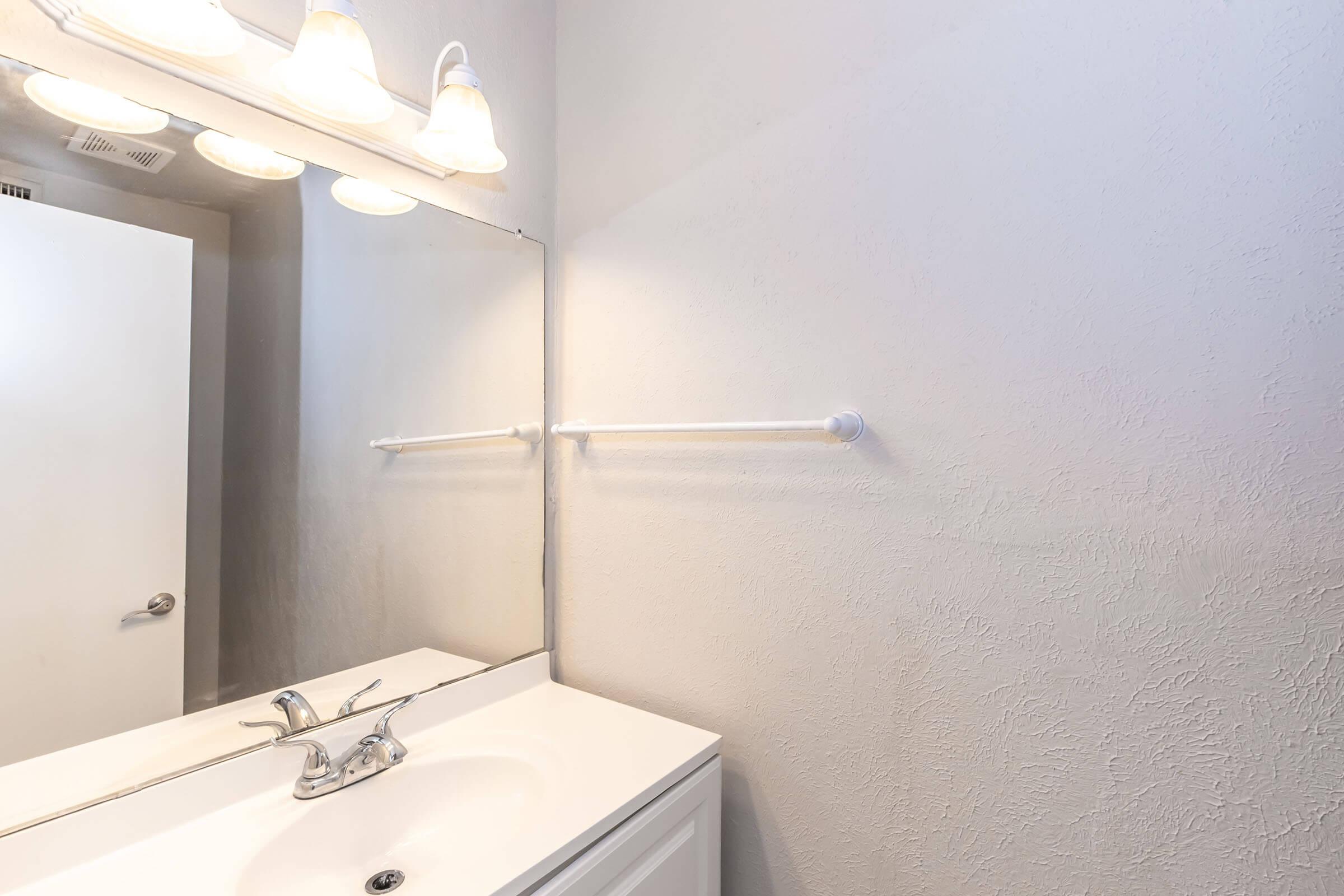 A bathroom sink area featuring a white countertop and a single faucet. Above the sink, there is a rectangular mirror and a light fixture with three lamps. A white towel bar is mounted on the wall next to the mirror, and a bathroom door is partially visible in the background. The walls are painted in a light color.