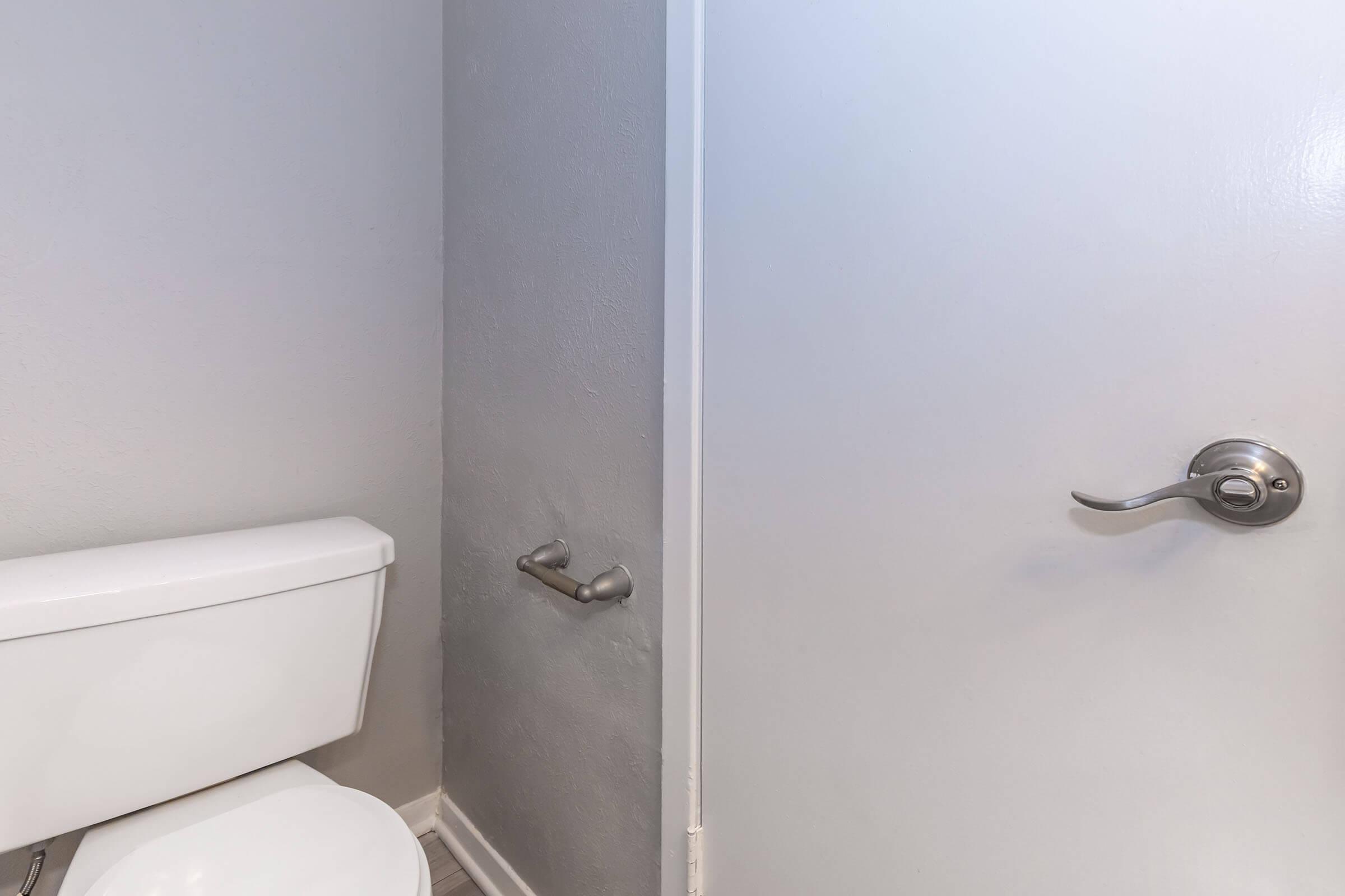 A small bathroom space featuring a white toilet next to a door. The wall is painted gray, and the door is closed with a shiny metal handle. A towel bar is mounted on the wall near the toilet. The overall atmosphere is simple and clean.