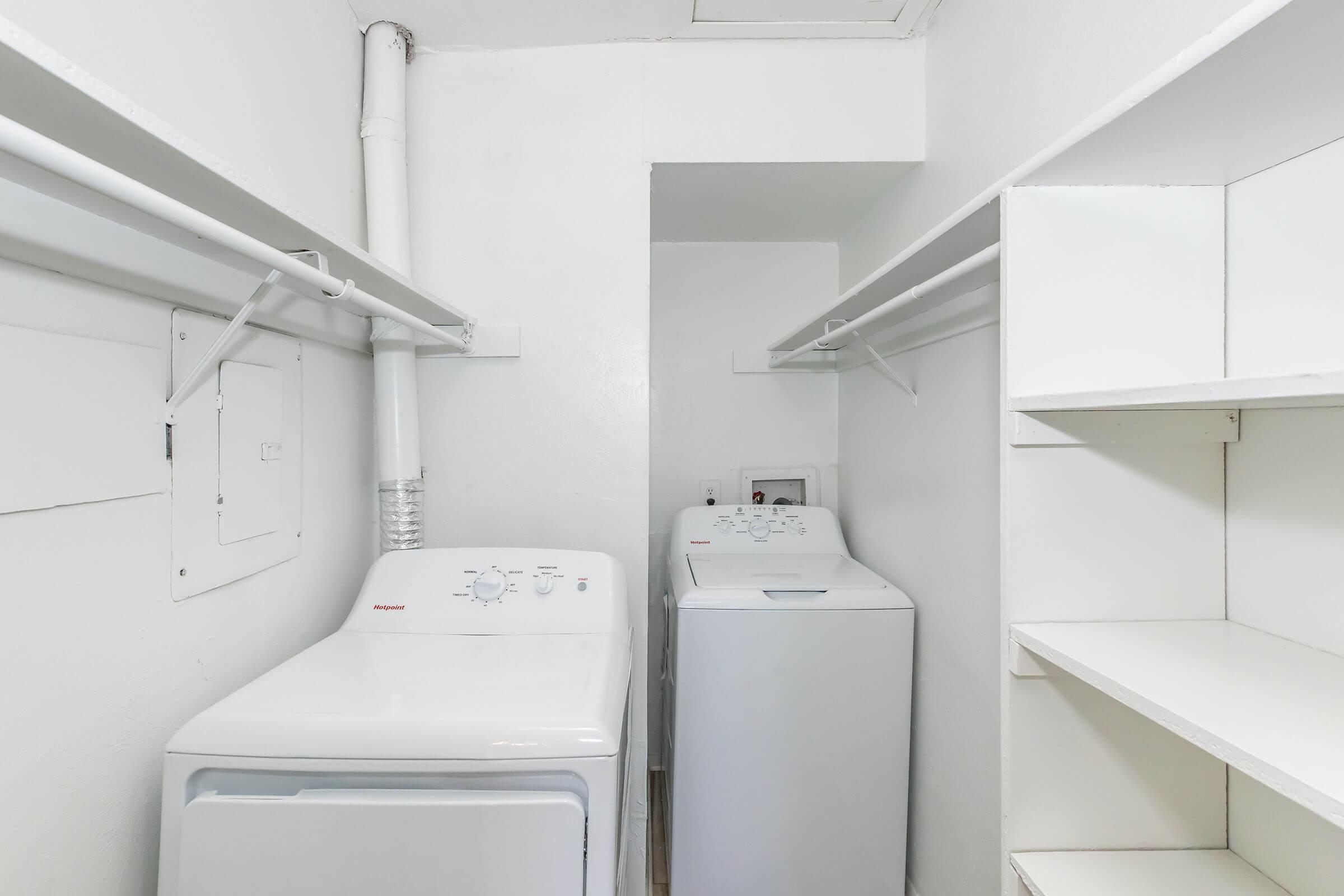 A clean and organized laundry room featuring a washer and dryer positioned side by side. The walls are painted white, and there are shelves for storage on the right side. The space is well-lit, with pipes visible on the ceiling.