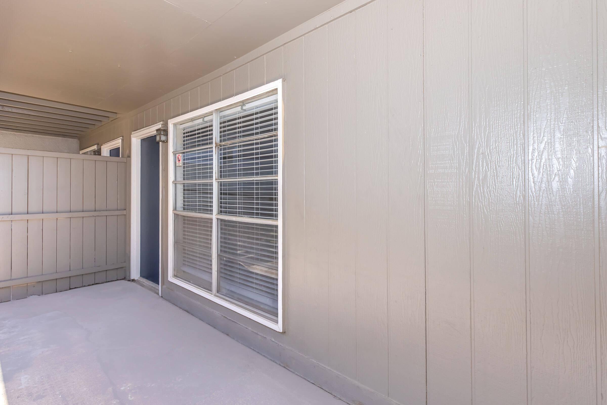 Exterior view of a grey building featuring a set of windows with a white frame, a door, and a fenced area. The surface appears smooth and painted, with a clean and minimalistic design, reflecting natural light. The setting is simple and uncluttered, focusing on the architectural elements.