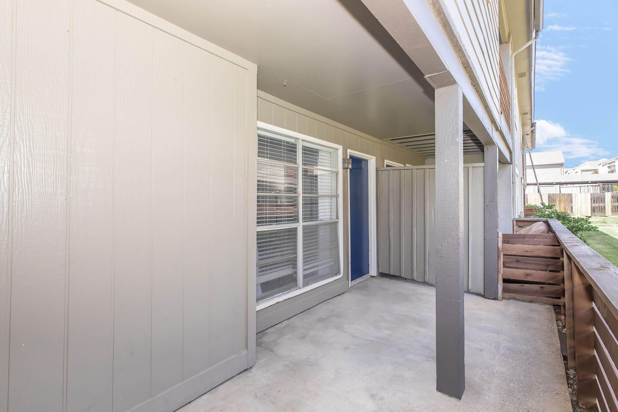 A covered patio area featuring a gray exterior with a blue door and large windows. The space is concrete-floored and includes wooden planters along one side. The surrounding area shows green grass and a clear sky, creating a bright and open atmosphere.