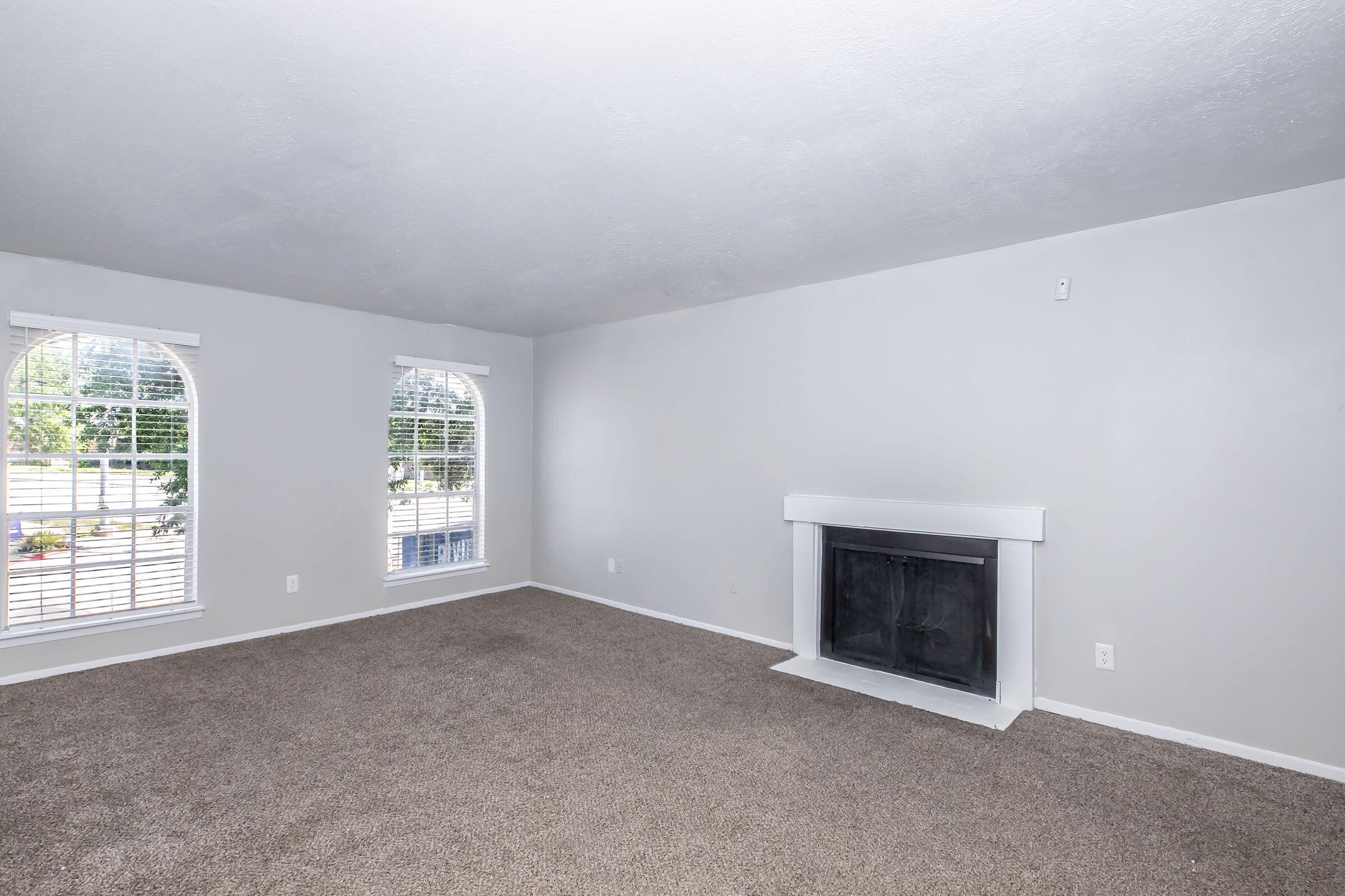Spacious living room with light gray walls and plush, beige carpet. Features two large arched windows with white blinds allowing natural light, and a simple white fireplace against the wall. The area is empty, creating an open and inviting atmosphere.