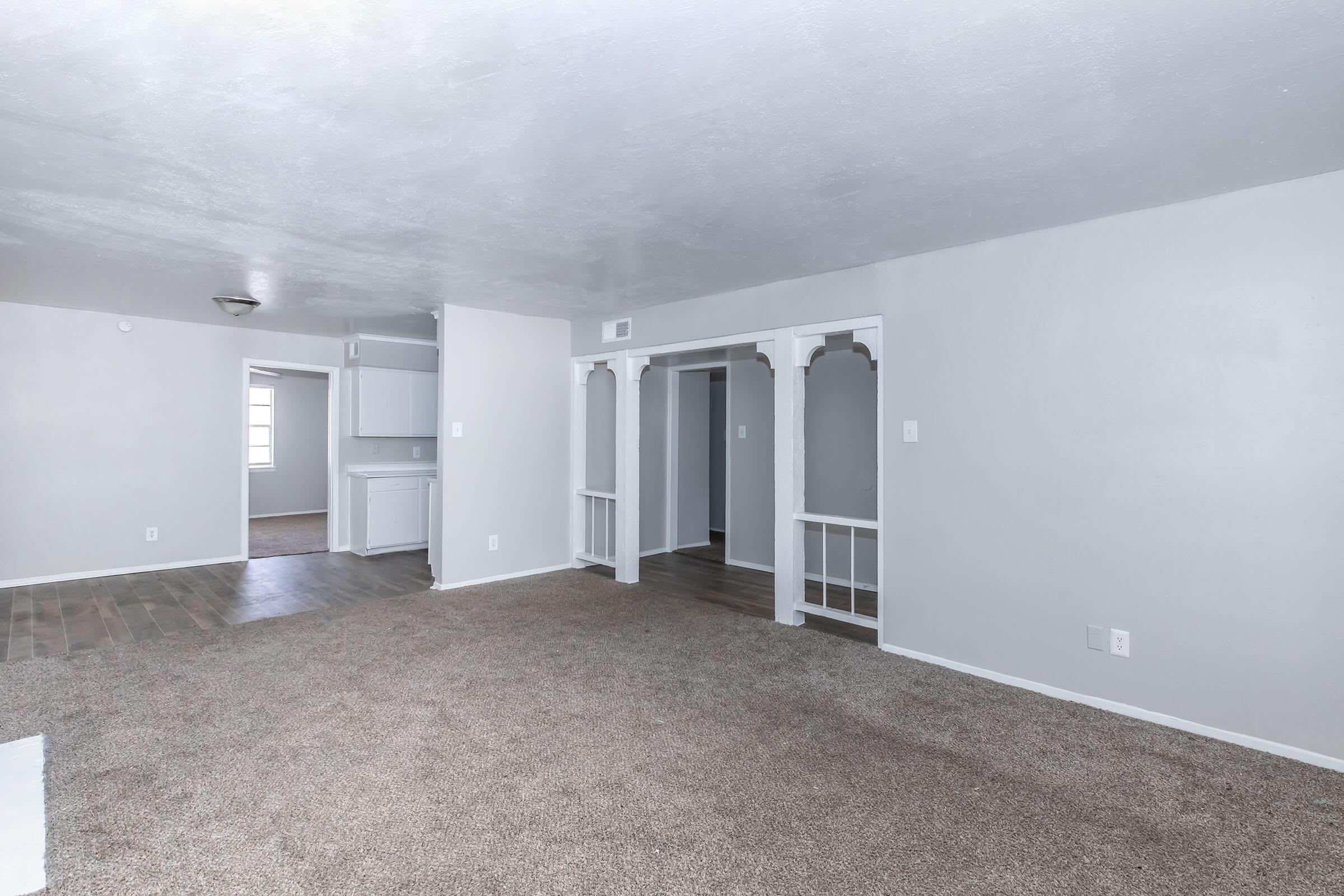 A spacious, airy living room with light gray walls and carpet, featuring a large, open layout. The room includes architectural details such as decorative columns, leading into a kitchen area with cabinets visible in the background. Natural light enters through a window.
