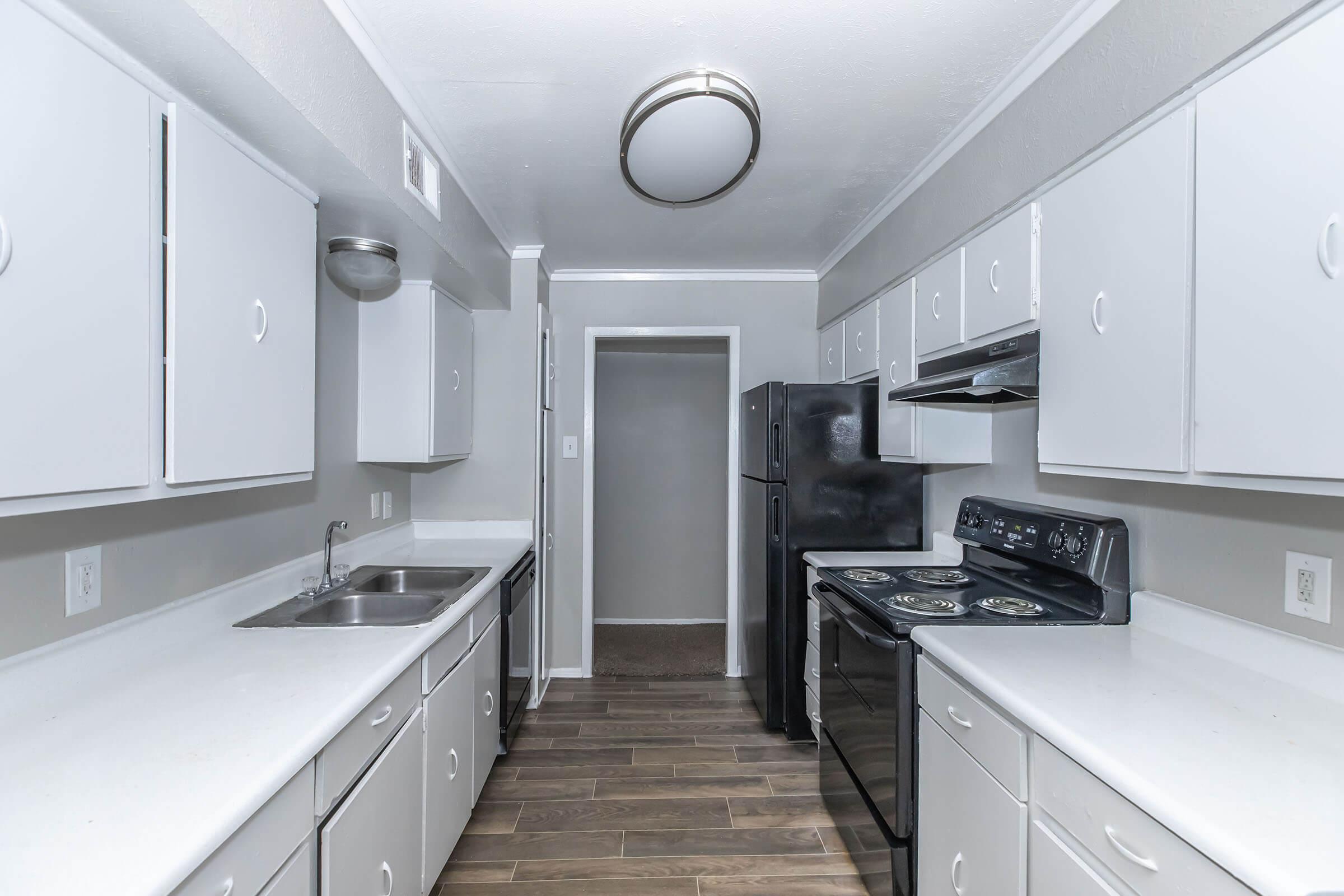 Modern kitchen with white cabinetry, a double sink, black appliances including a refrigerator and stove, and a light fixture on the ceiling. The floor features wooden tiles, and there is an open doorway leading to another room. The overall design is clean and contemporary.