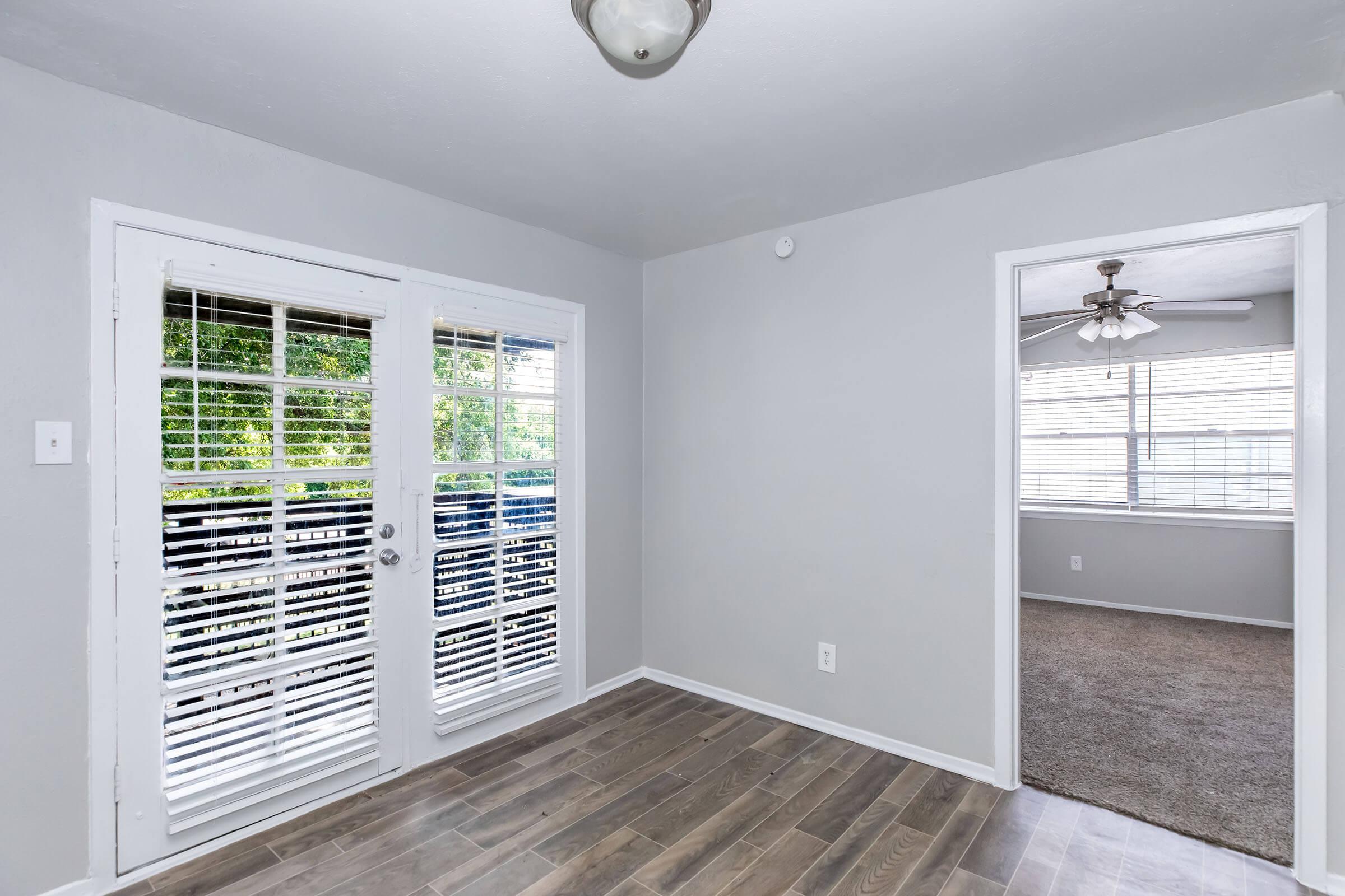 Interior view of a light gray room featuring a set of double doors with white shutters leading to a balcony. The floor is made of wood. To the right, there's an open doorway into another room with a ceiling fan and large windows with blinds. Walls are painted a soft gray.