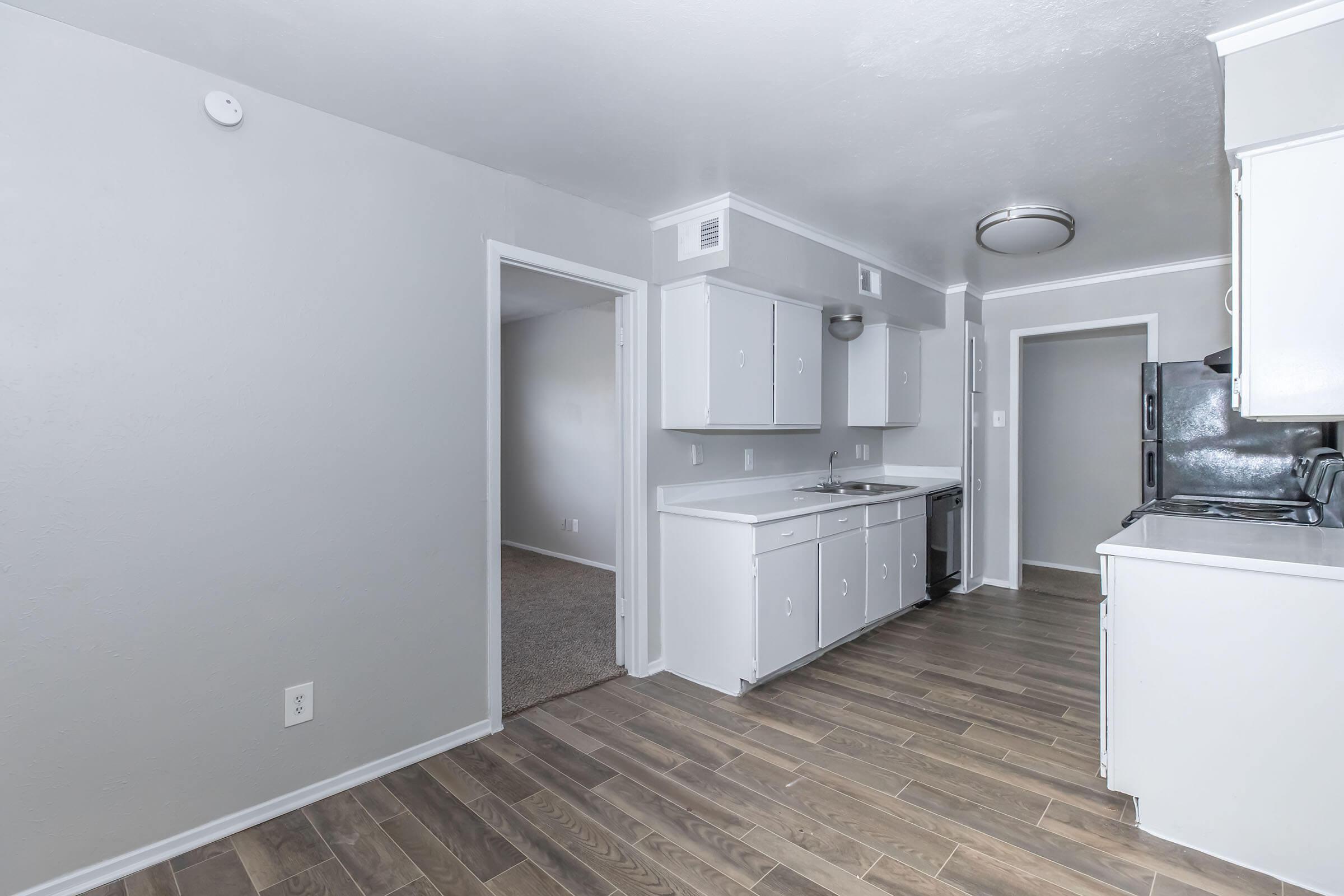A modern kitchen featuring white cabinets, stainless steel appliances, and a tile floor. The open layout includes a doorway leading to a carpeted living area and another room. The walls are painted in soft gray tones, enhancing the bright and airy feel of the space.