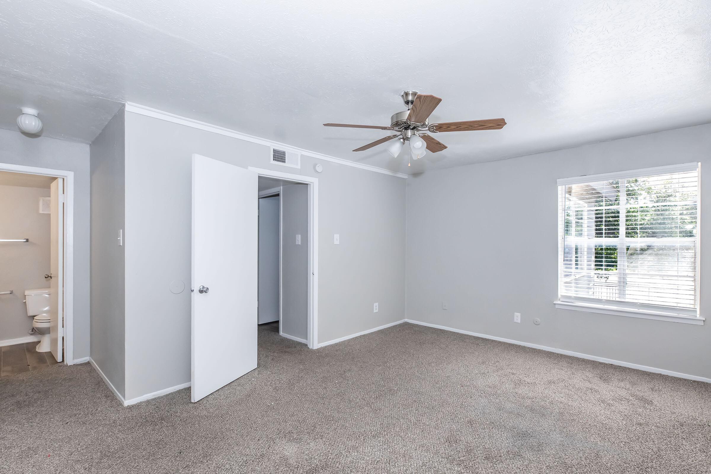 A spacious, empty room with light gray walls and carpet. Features include a ceiling fan, a window with blinds allowing natural light, and a doorway leading to a bathroom. The overall ambiance is clean and well-lit, suitable for various uses.
