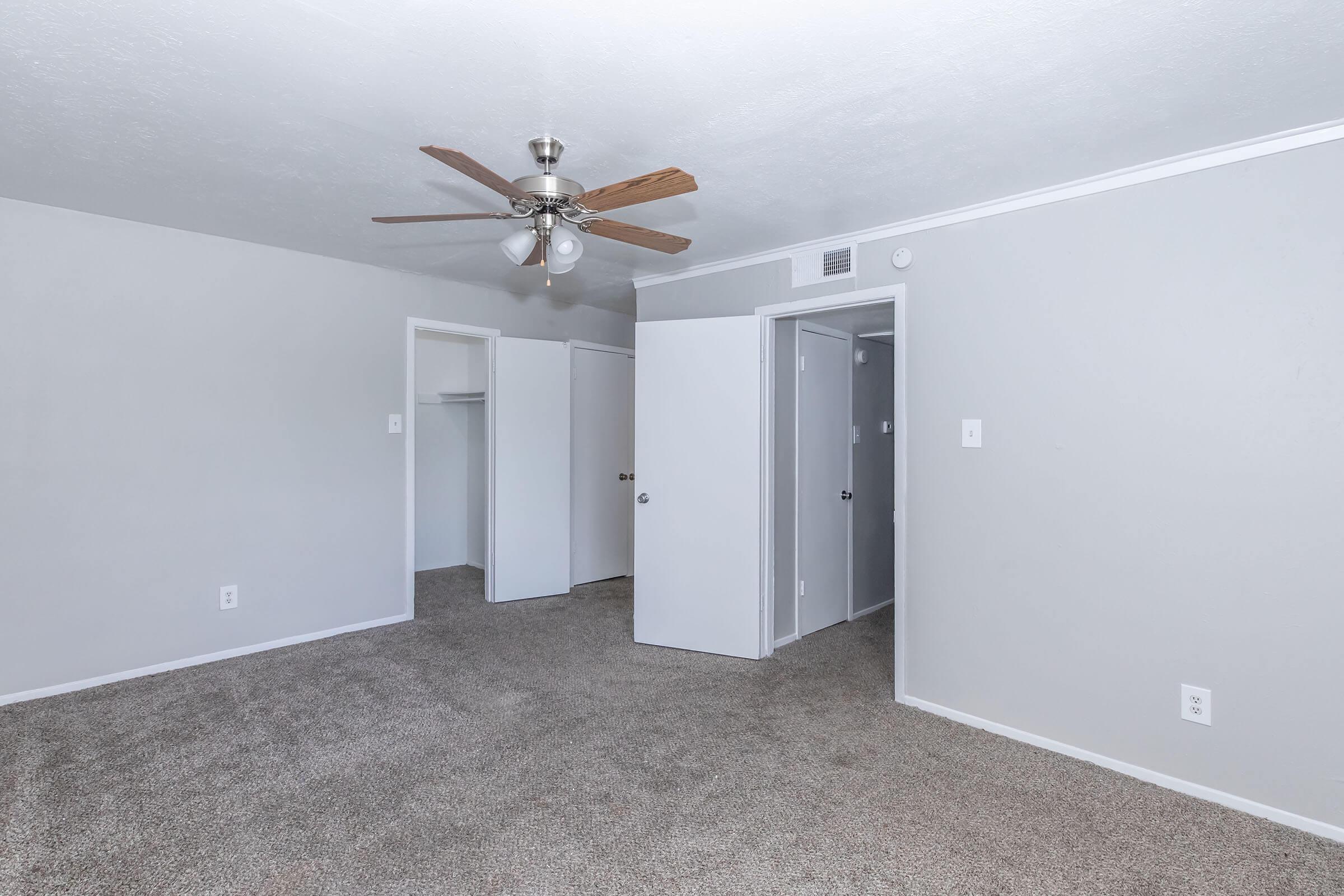 Spacious bedroom with light gray walls and beige carpet flooring. A ceiling fan is mounted in the center, and there are two doors leading to closets on the left. The room has a clean and modern appearance, featuring a bright, open layout. Natural light may enter through a hidden window.
