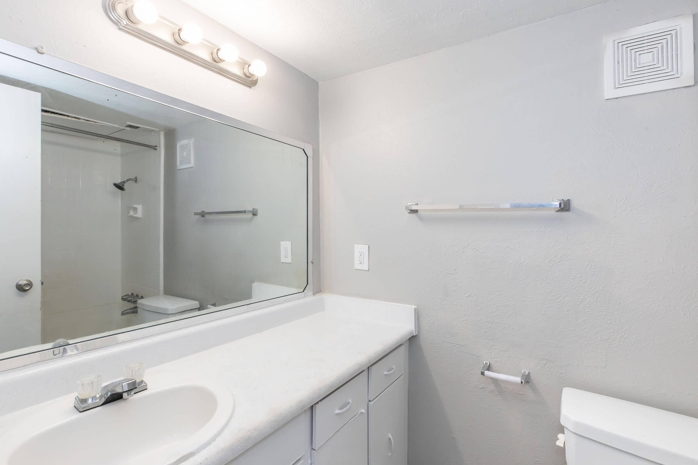 A clean bathroom with a light gray wall color. It features a large mirror above a white countertop with sink and faucet. There is a toilet visible in the corner, along with a towel bar and a small shower area. Bright lighting is provided by a row of light bulbs above the mirror.