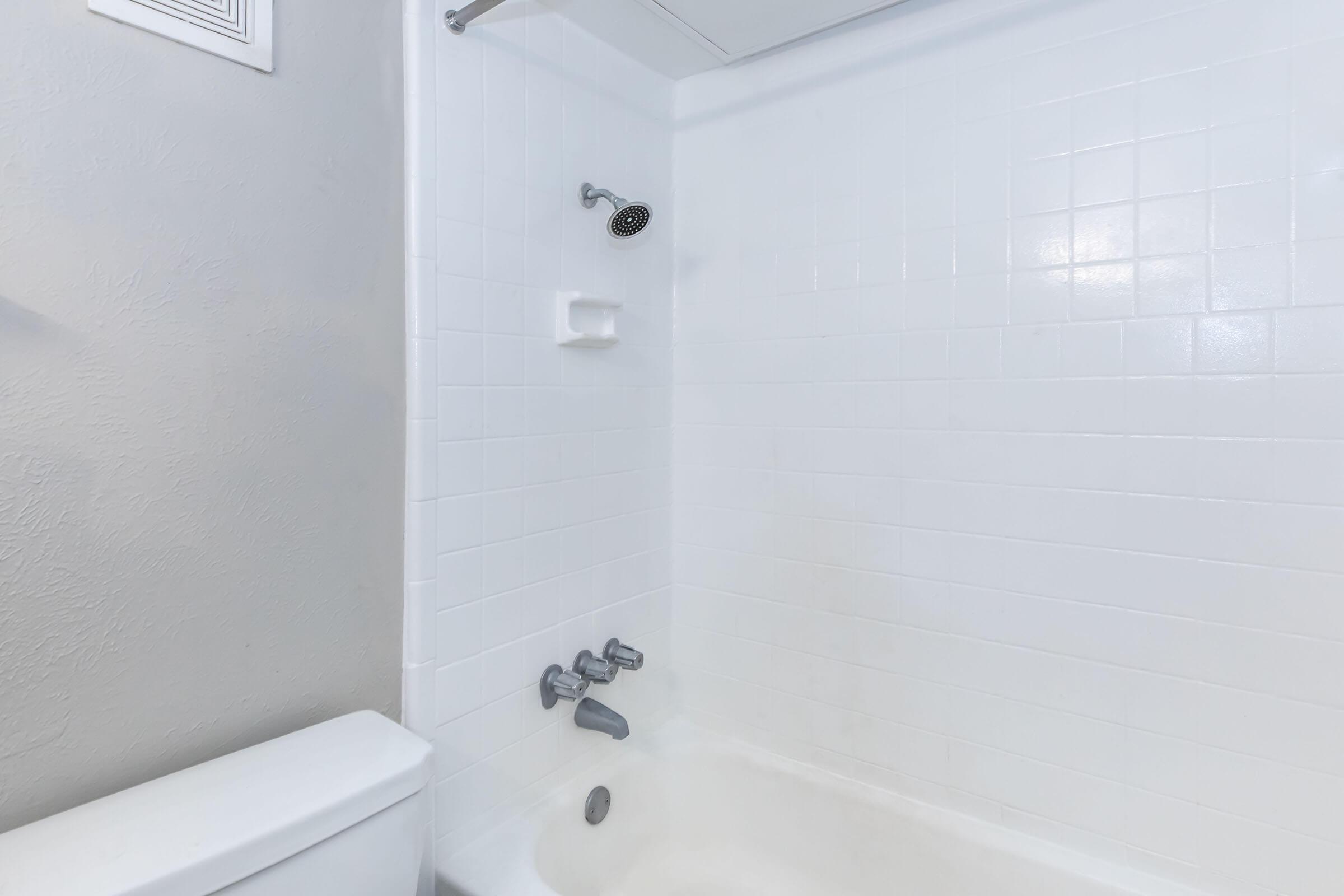 A clean, modern bathroom featuring a white tiled shower area with a showerhead, a built-in soap dish, and a bathtub. The toilet is visible to the left, with neutral-colored walls that enhance the bright and airy feel of the space.