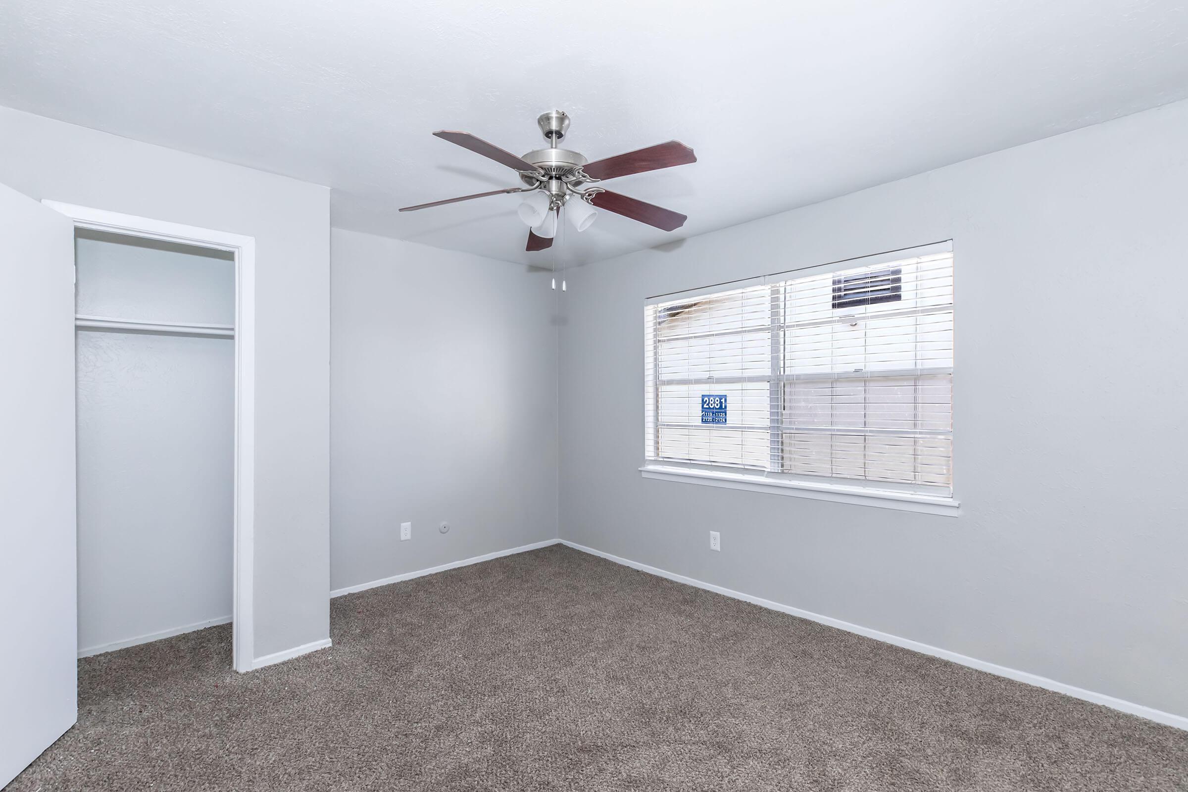 Empty bedroom featuring light gray walls and carpet. A ceiling fan with dark wood blades is mounted above. There is a window with blinds allowing natural light and a closet with a white door on the left. The room appears clean and spacious, suitable for furniture arrangement.