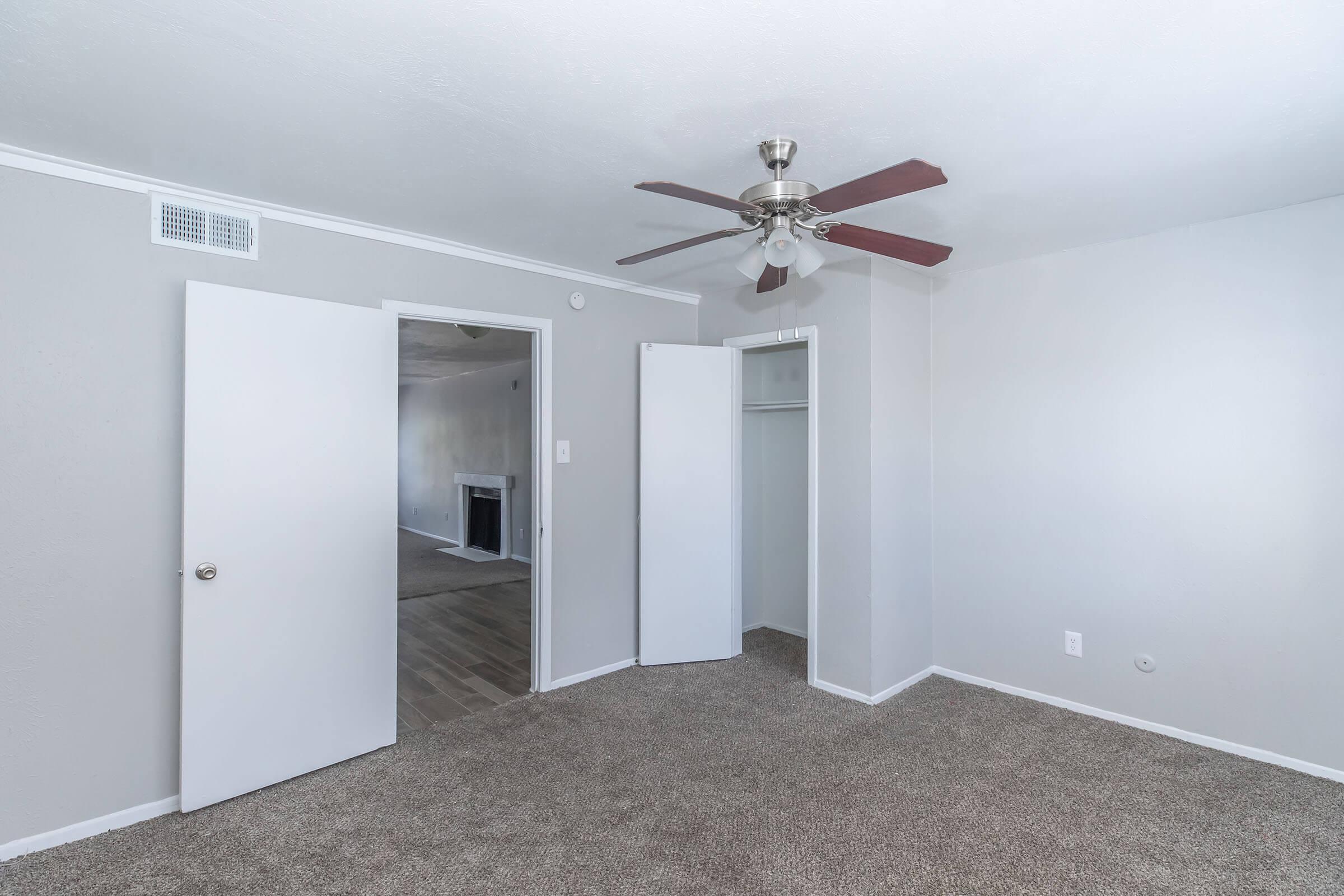 A spacious, light gray room with carpeted flooring, featuring a ceiling fan and two closed white doors on the left. One door leads to a closet. In the background, a doorway opens to another room, possibly a living area, with a fireplace visible. Natural light brightens the space.