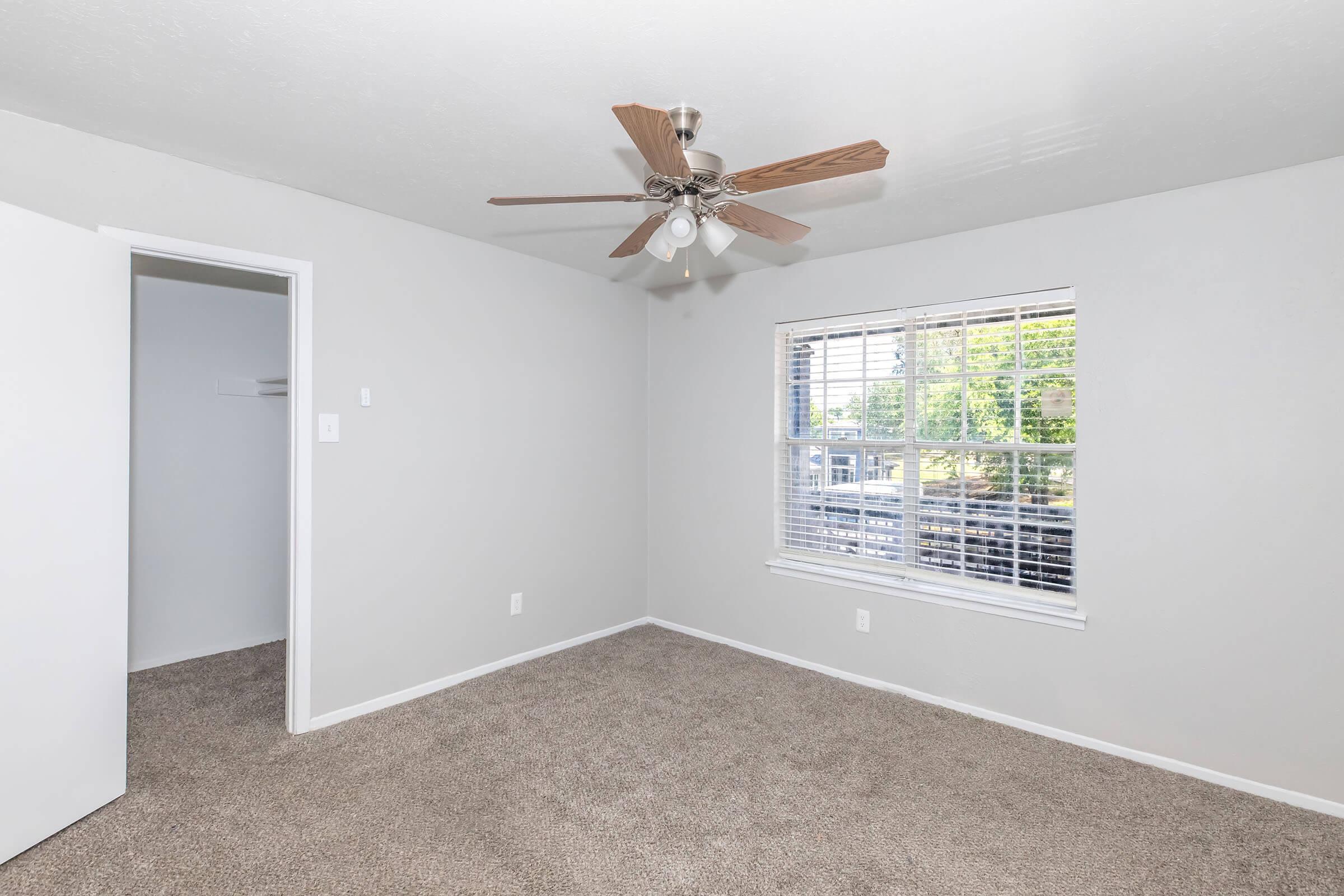 A light and airy bedroom featuring beige carpet, a ceiling fan with wooden blades, and a large window with blinds. The room has light gray walls and a closet with a white door, creating a clean and modern aesthetic. Natural light brightens the space, enhancing its inviting atmosphere.