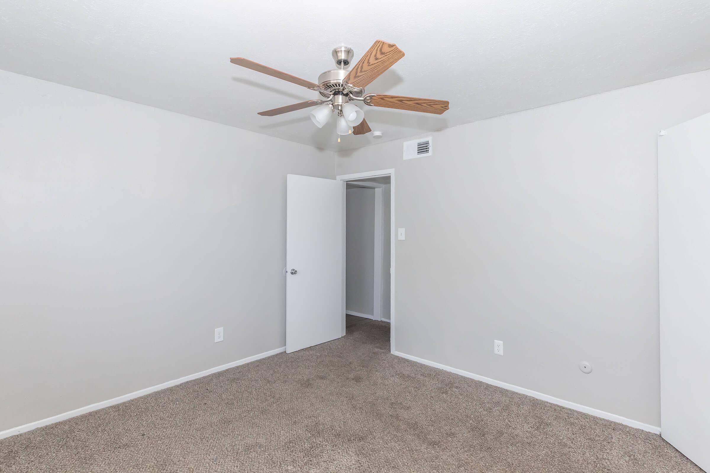A light and airy bedroom with beige carpet and light gray walls. The room features a ceiling fan with wooden blades and a door leading to a small closet. Another door opens to a hallway. The space is empty, creating a fresh, inviting atmosphere.