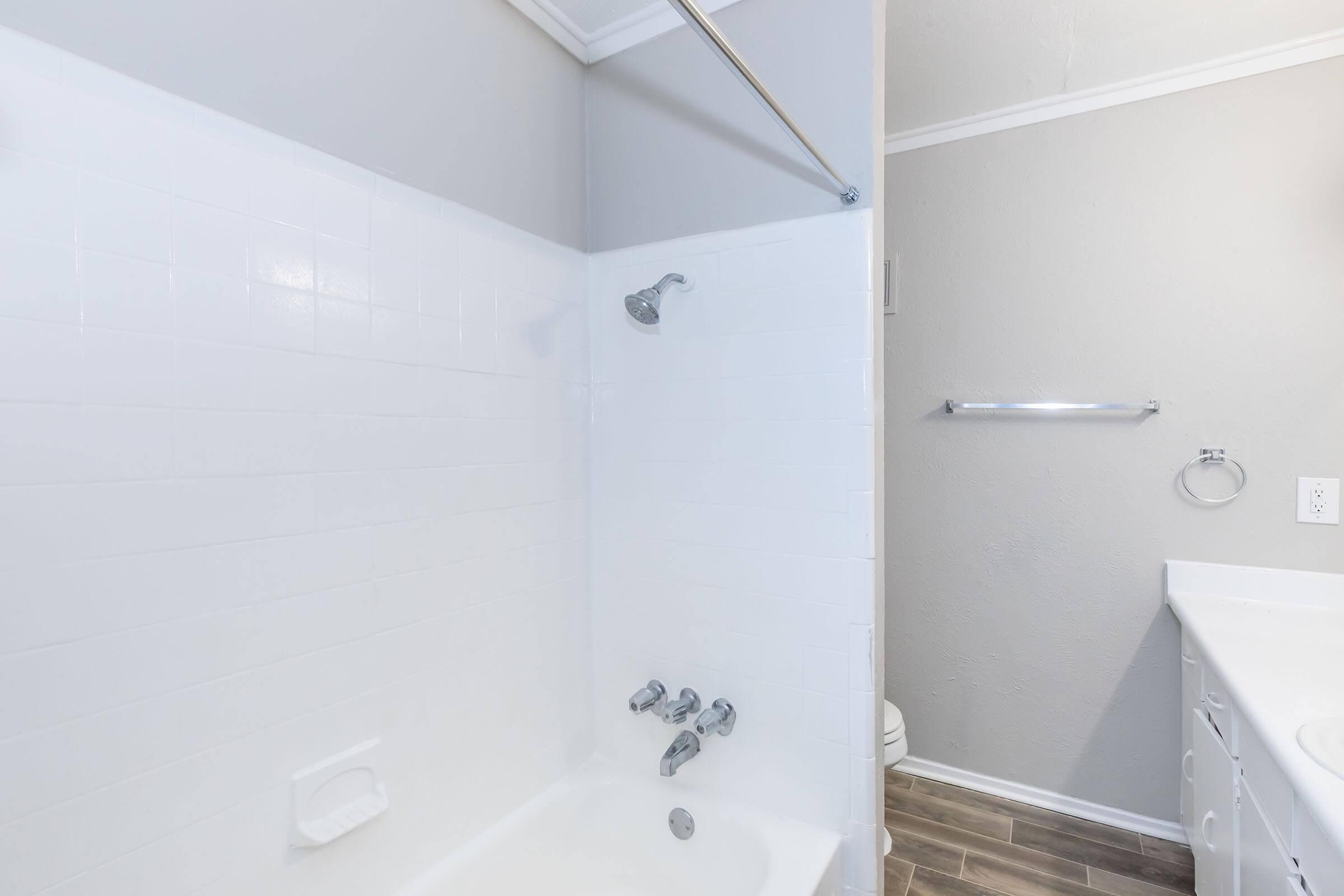 A clean, modern bathroom featuring a white tiled shower and bathtub combination. The bathtub has three faucet controls, and there's a showerhead mounted above. Additionally, a towel rack and a ring for hanging towels are visible, set against light gray walls, with a light-colored countertop in the background.