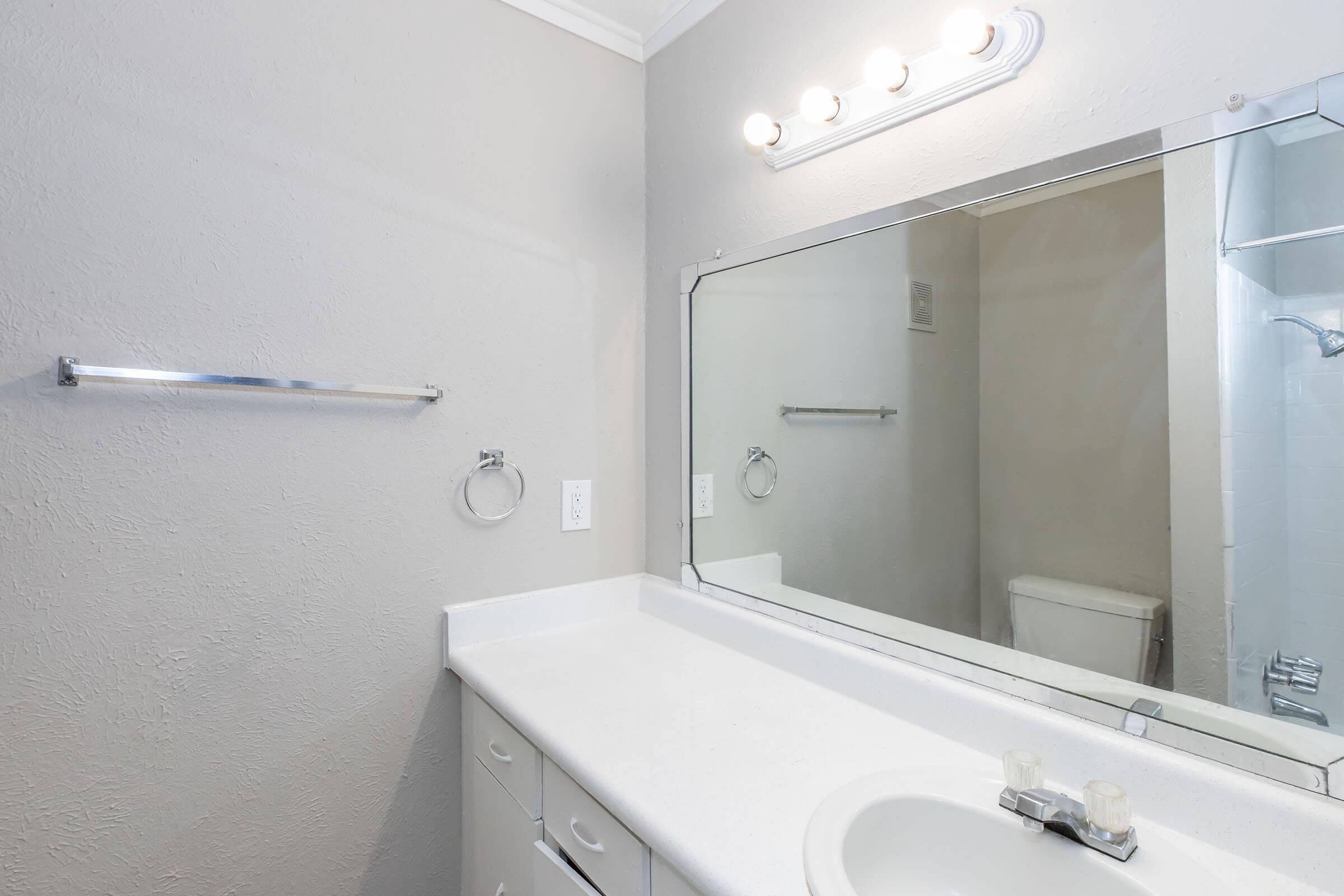 A clean, modern bathroom with a white countertop and sink, a large mirror above the sink, a towel rack on the wall, and a separate shower area visible in the background. The walls are painted in a light gray color, and there is a bright overhead light fixture.