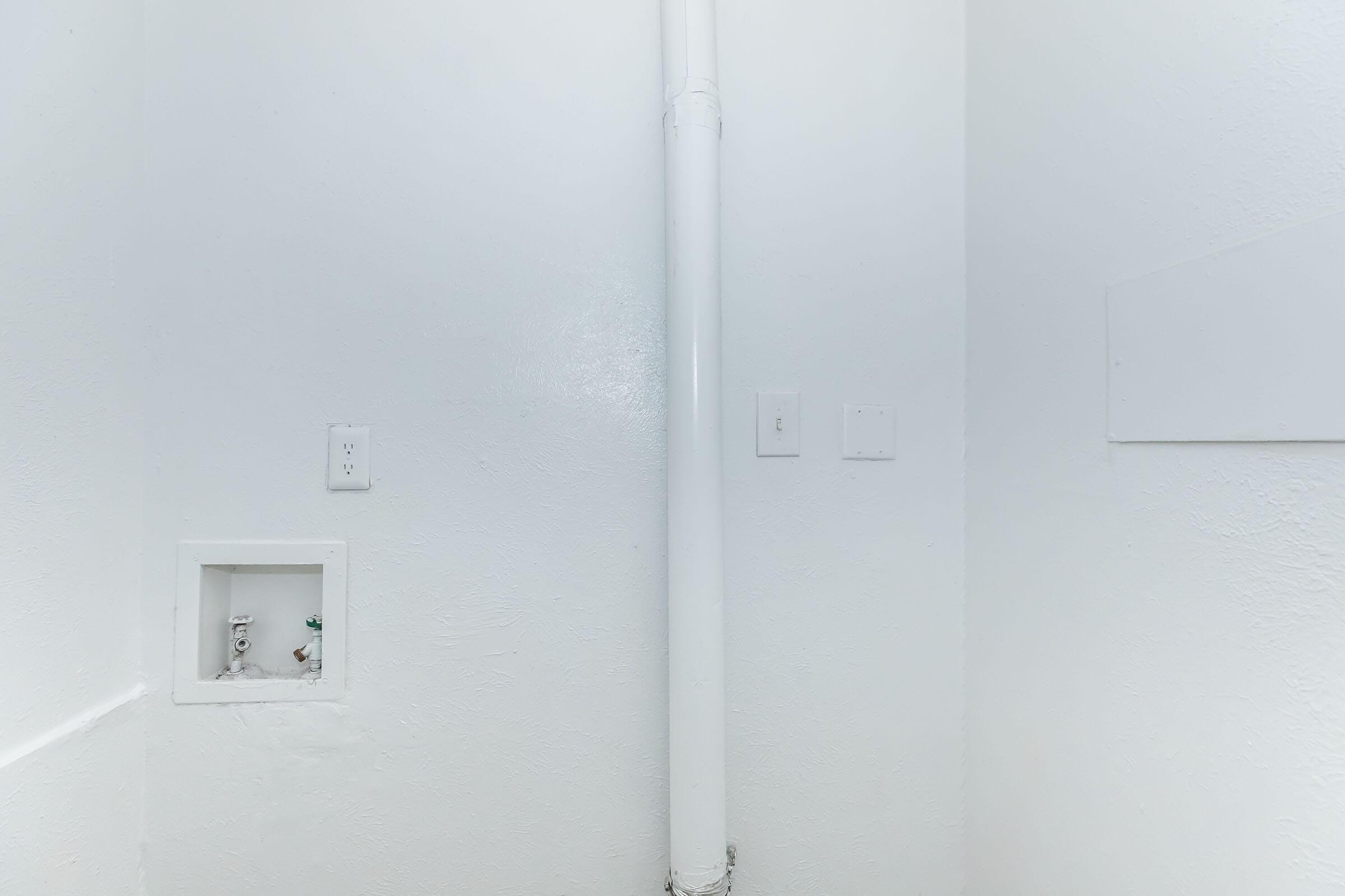 A clean, white-walled corner of a room featuring a vertical white pipe running along the wall. There are two electrical outlets, and a small, recessed area in the wall that appears to have plumbing connections. The overall setting is minimalist and well-lit.