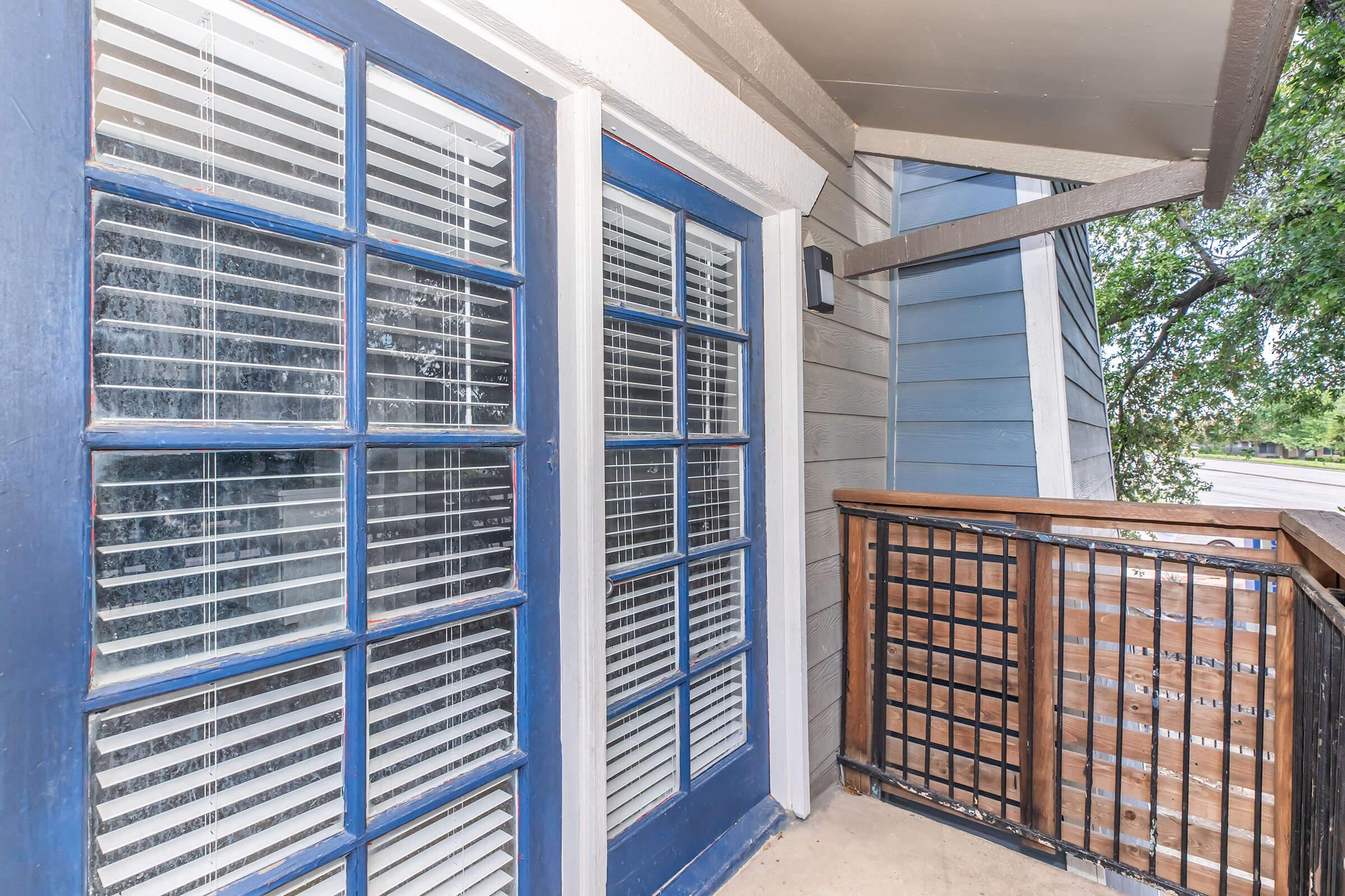 View of a balcony with wooden railing and large windows featuring blue frames. The windows have white blinds, and the surrounding building features gray siding. There are trees visible outside the balcony.