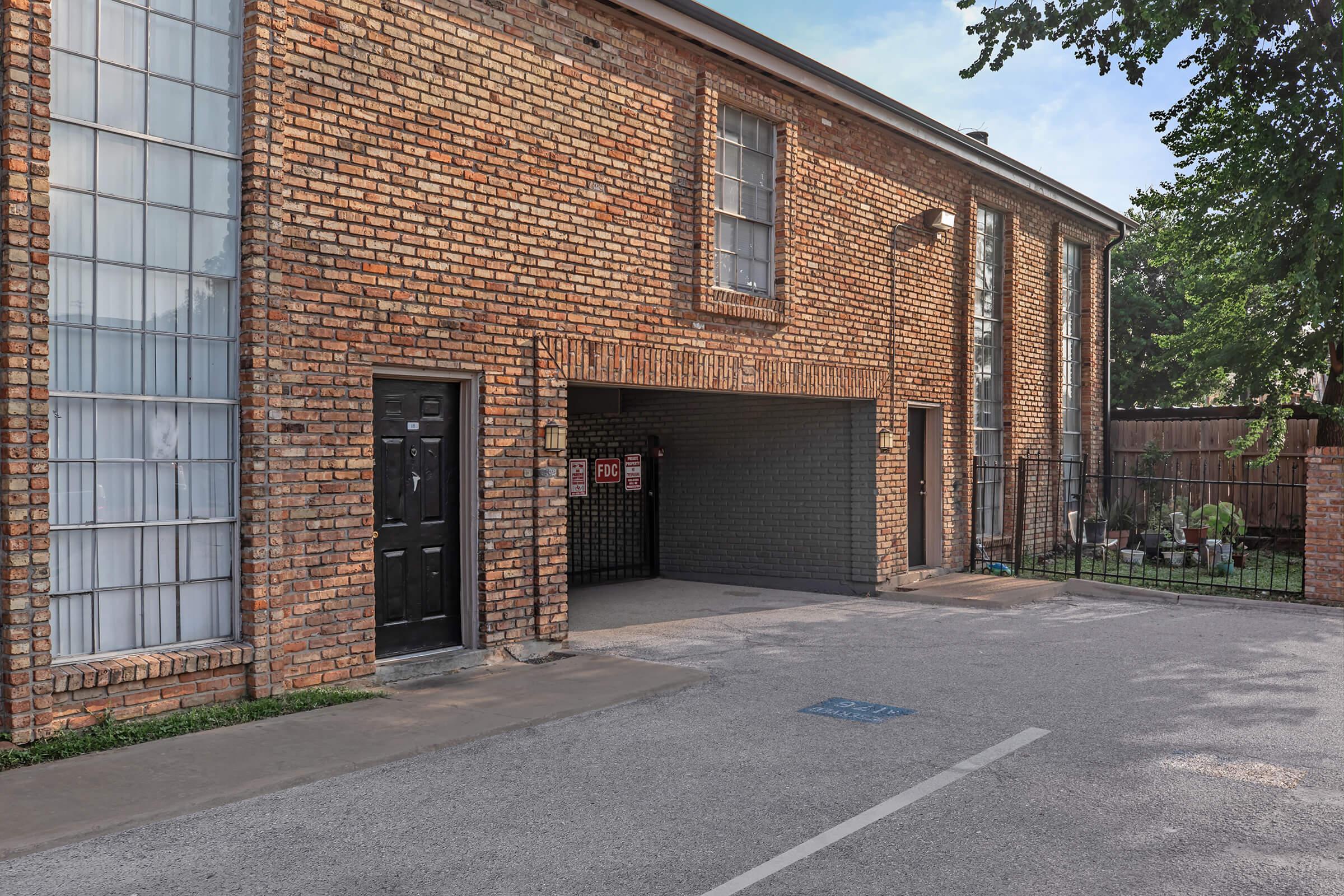 A brick building featuring large windows and a black door. An open garage area is visible, with a sign hanging that reads "For Rent." The scene includes a paved parking area and surrounding greenery, providing a clear view of the structure's exterior and its entrance.