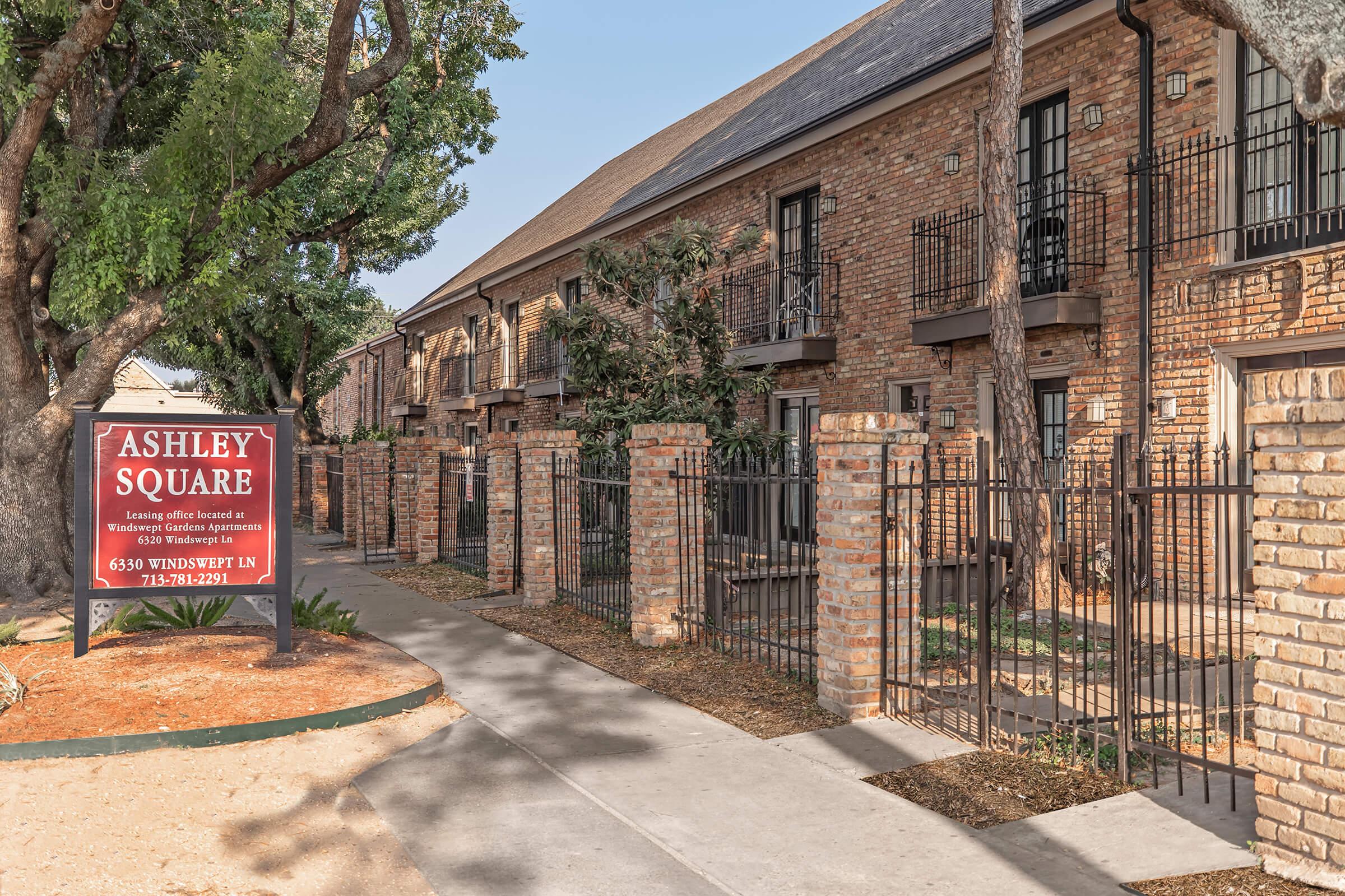 A brick apartment complex named "Ashley Square," featuring multiple units with balconies. The entrance is framed by a decorative fence and landscaping, with a prominent sign displaying the name and contact information. The pathway leads to the building, surrounded by trees.