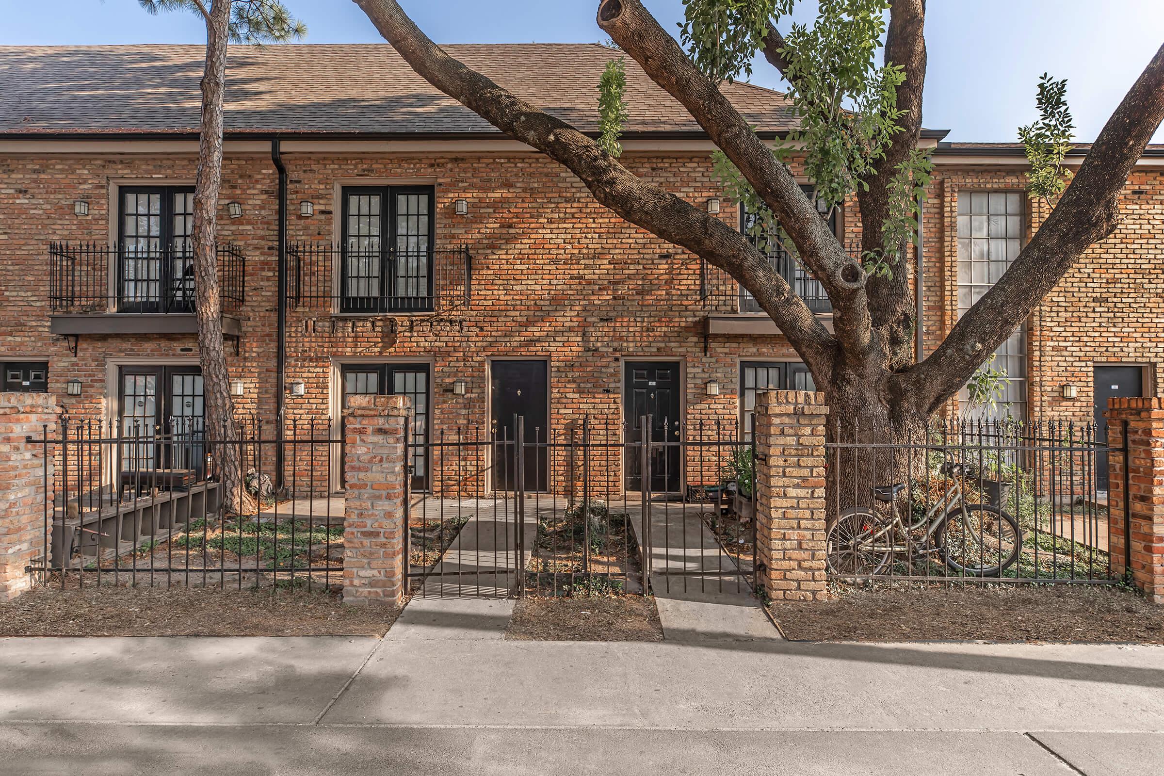 A brick building with multiple units, each featuring a front porch and black doors. A large tree grows near the building, and there are bike racks in front. A paved walkway leads up to the entrance, and the area is enclosed by a wrought iron fence. The setting is urban and well-maintained.