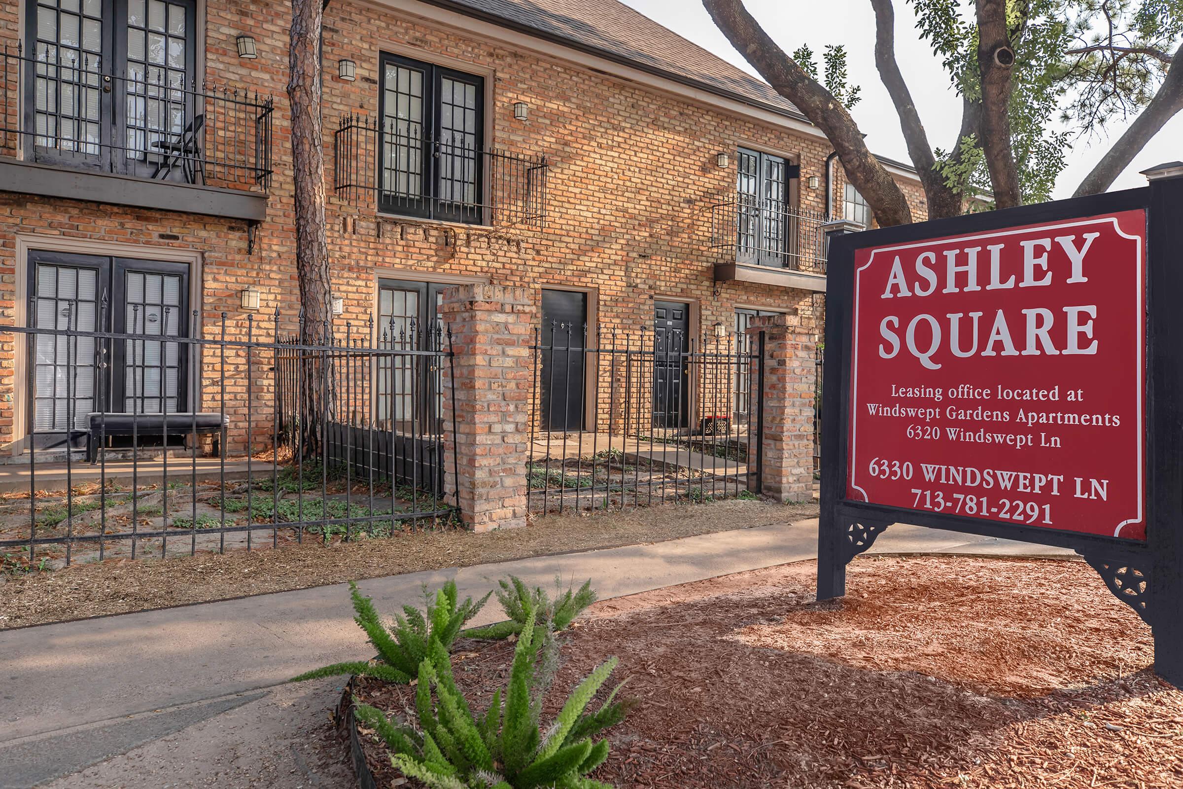 A brick apartment building with multiple units featuring balconies. In the foreground, a sign displays "Ashley Square" with leasing information, surrounded by landscaping, including shrubs. The setting includes a pathway leading to the entrance.