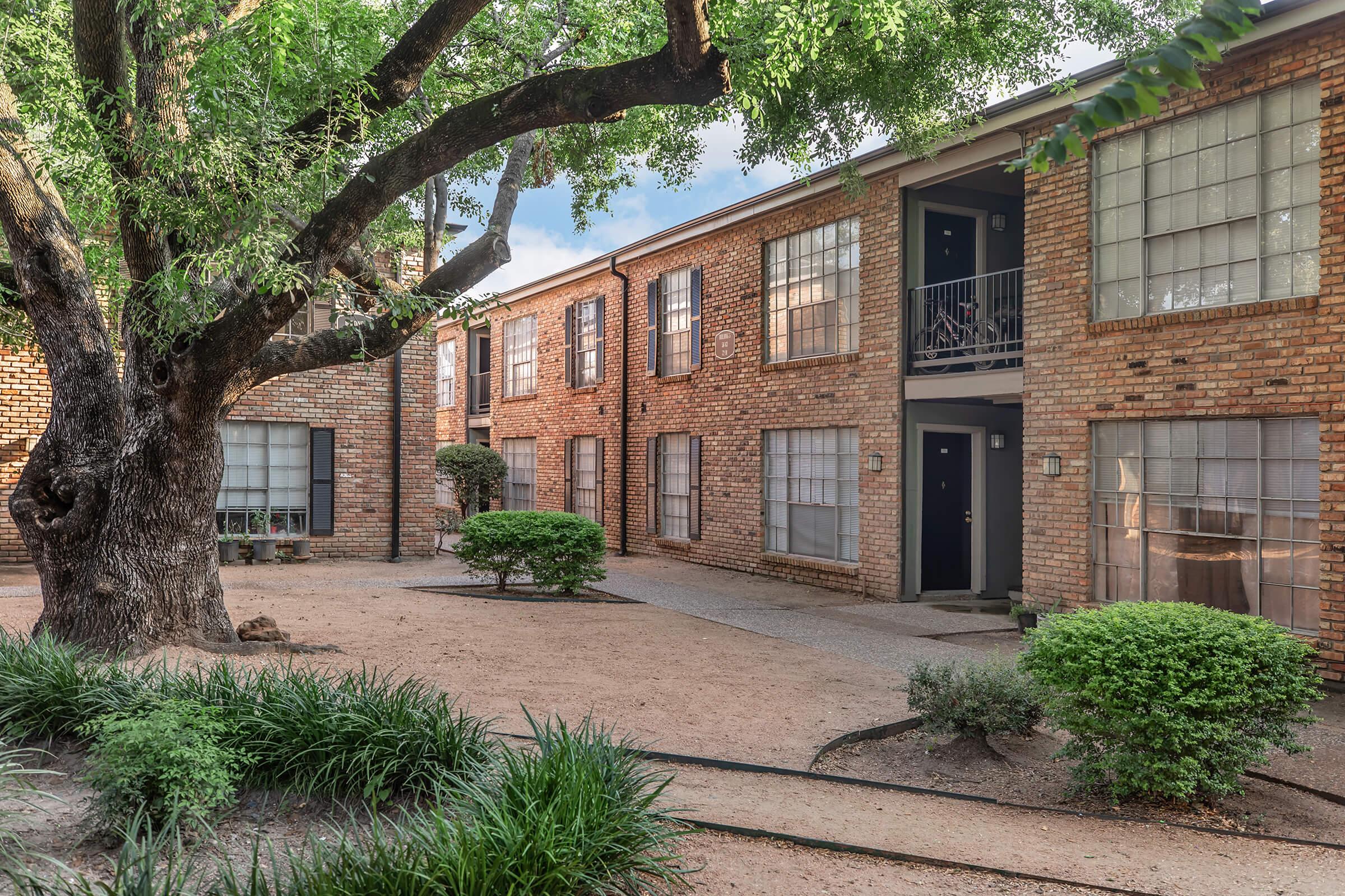 A peaceful courtyard featuring brick buildings with multiple windows, surrounded by well-maintained greenery including shrubs and grass. A large tree provides shade in the center, creating a relaxing outdoor space. The scene is sunny and inviting, highlighting the charm of this residential area.