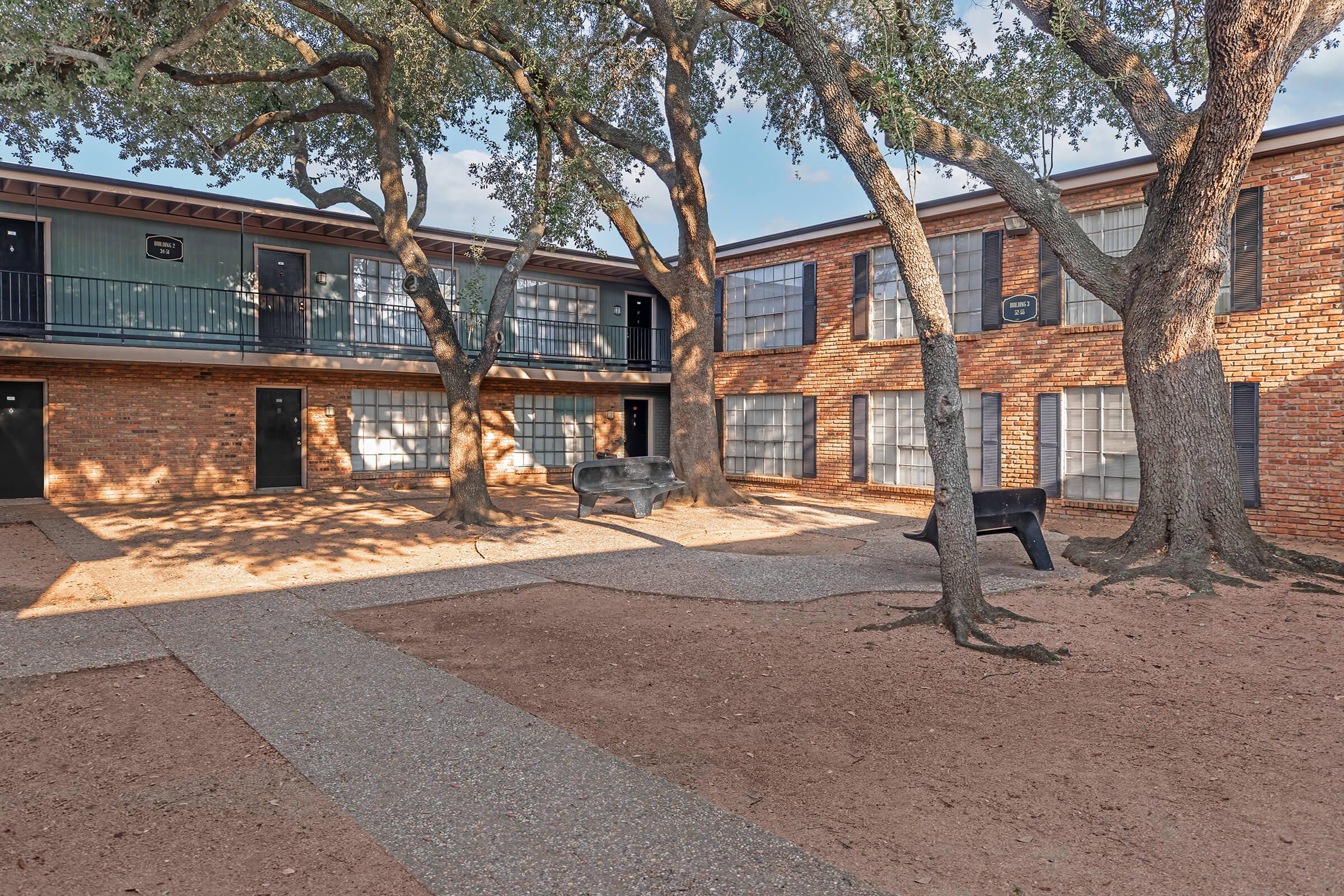 View of a shaded courtyard between two brick apartment buildings, featuring several trees, a gravel pathway, and outdoor seating. Sunlight filters through the branches, creating dappled patterns on the ground. The atmosphere is tranquil and inviting.