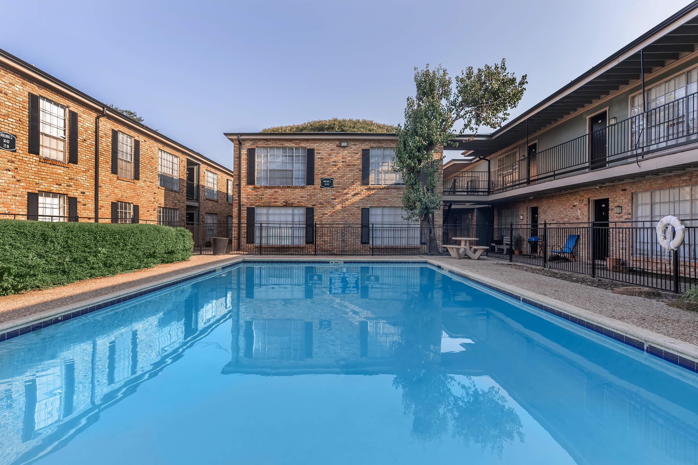 A clear blue swimming pool at an apartment complex, surrounded by brick buildings. Lush greenery and a tree are visible near the pool area. The buildings have balconies, and a picnic table is positioned by the poolside, creating a relaxing outdoor space. The sky is partly sunny, adding to the inviting atmosphere.