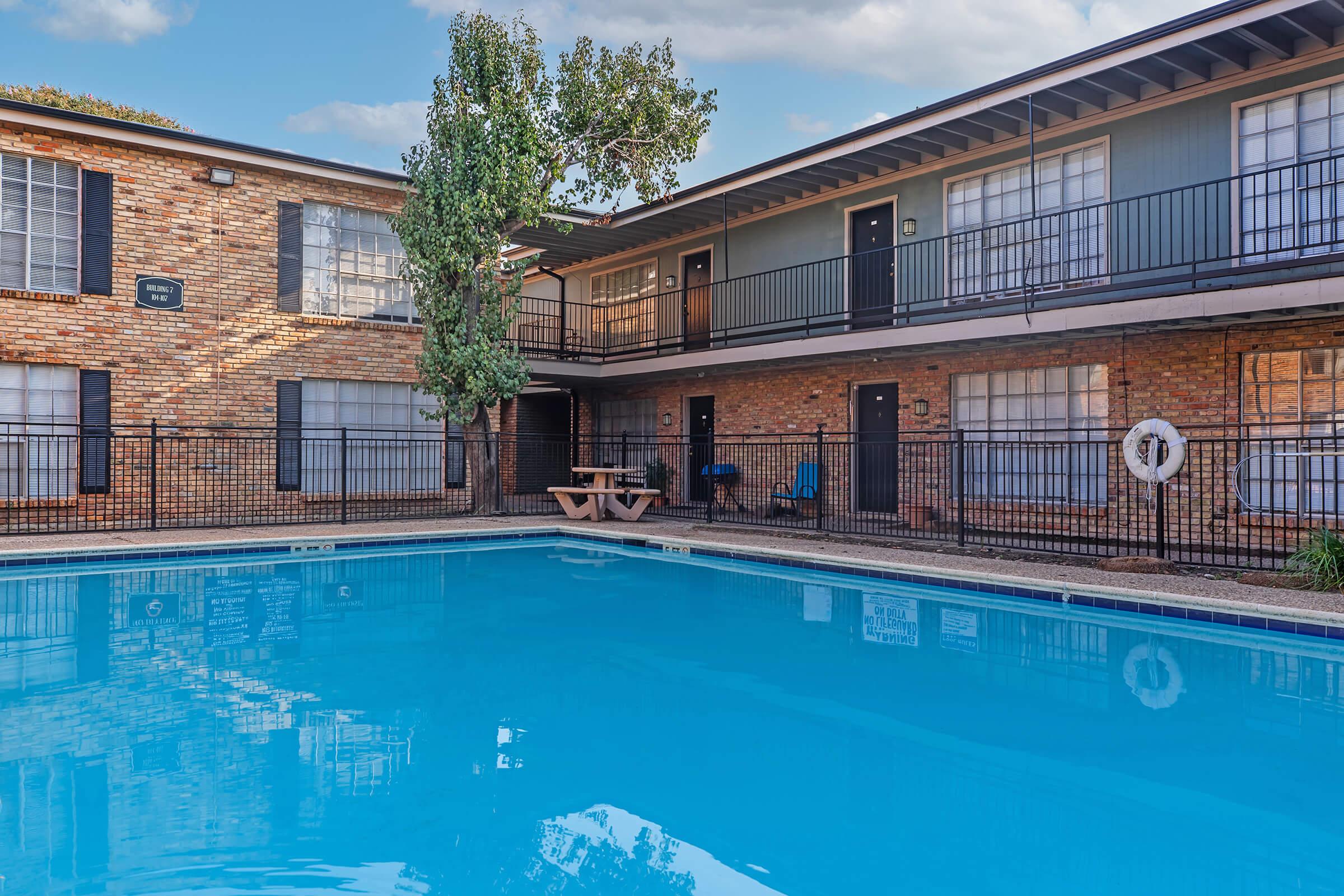 A clear blue swimming pool surrounded by a brick building with multiple balconies. There are picnic tables and chairs near the pool area, and a few trees providing some shade. The sky is partly cloudy, creating a bright and inviting atmosphere.