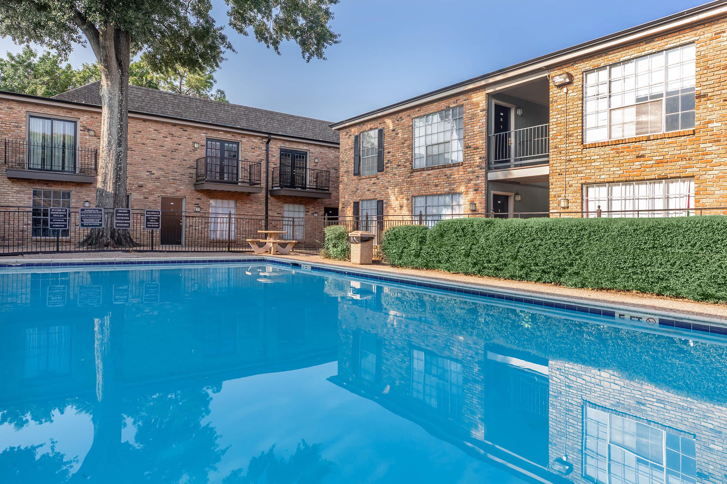 A clear blue swimming pool surrounded by greenery and an apartment complex with brick exteriors. The pool reflects the building and trees nearby, creating a tranquil atmosphere. The area features outdoor seating by the pool and balconies on the second floor of the apartments.