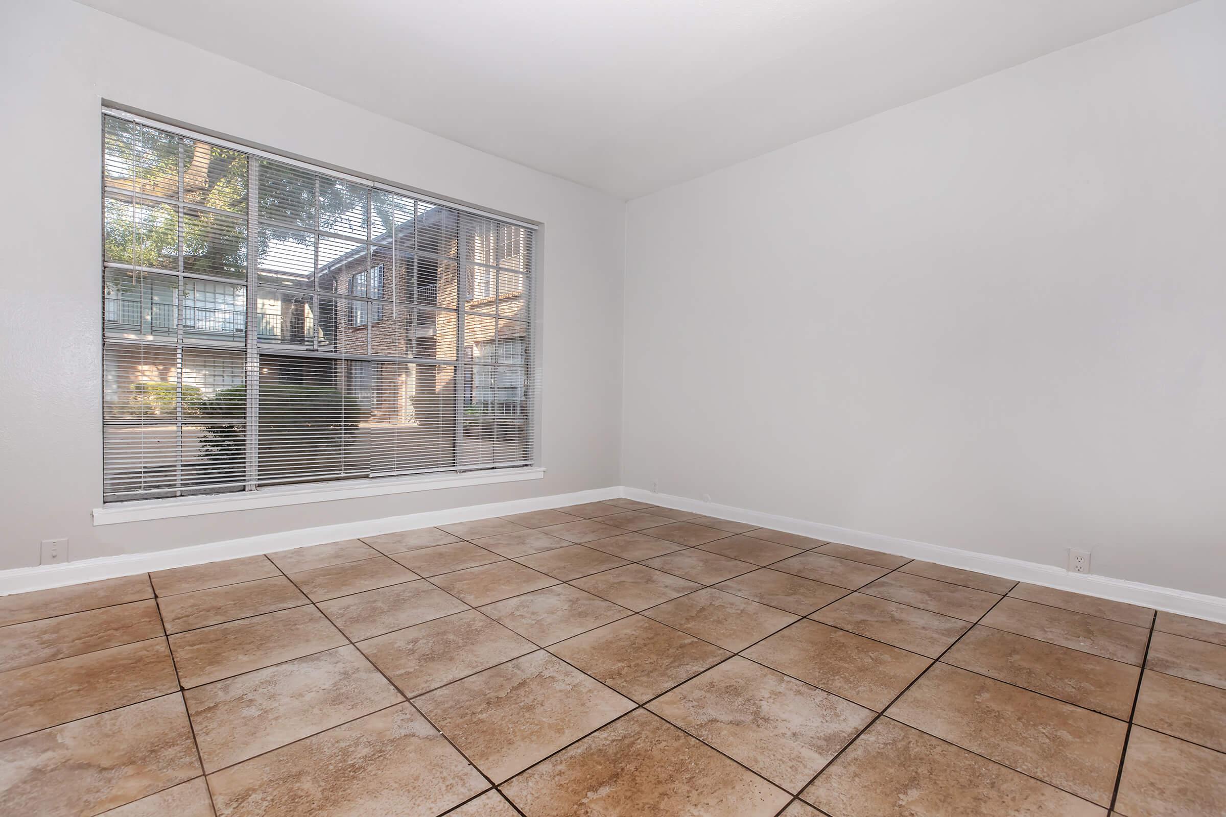 Empty room with beige tiled flooring and a large window allowing natural light. The window features blinds and overlooks greenery outside. The walls are painted white, creating a bright and airy atmosphere. No furniture or decorations are present, giving the space a minimalistic look.