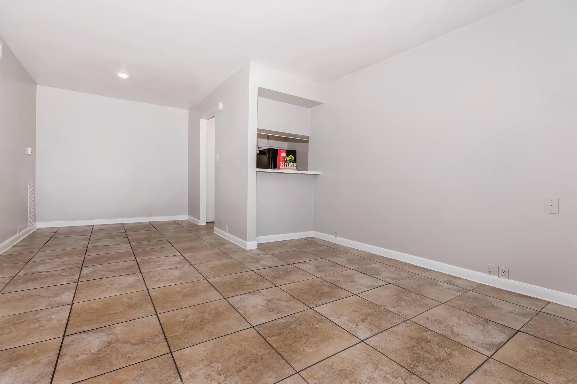 Empty room with light gray walls and a tiled floor. There is a small open kitchen area with a bar counter on one side. A microwave is visible on the counter, and the overall space is well-lit with natural light. The room is spacious and ready for furnishing.