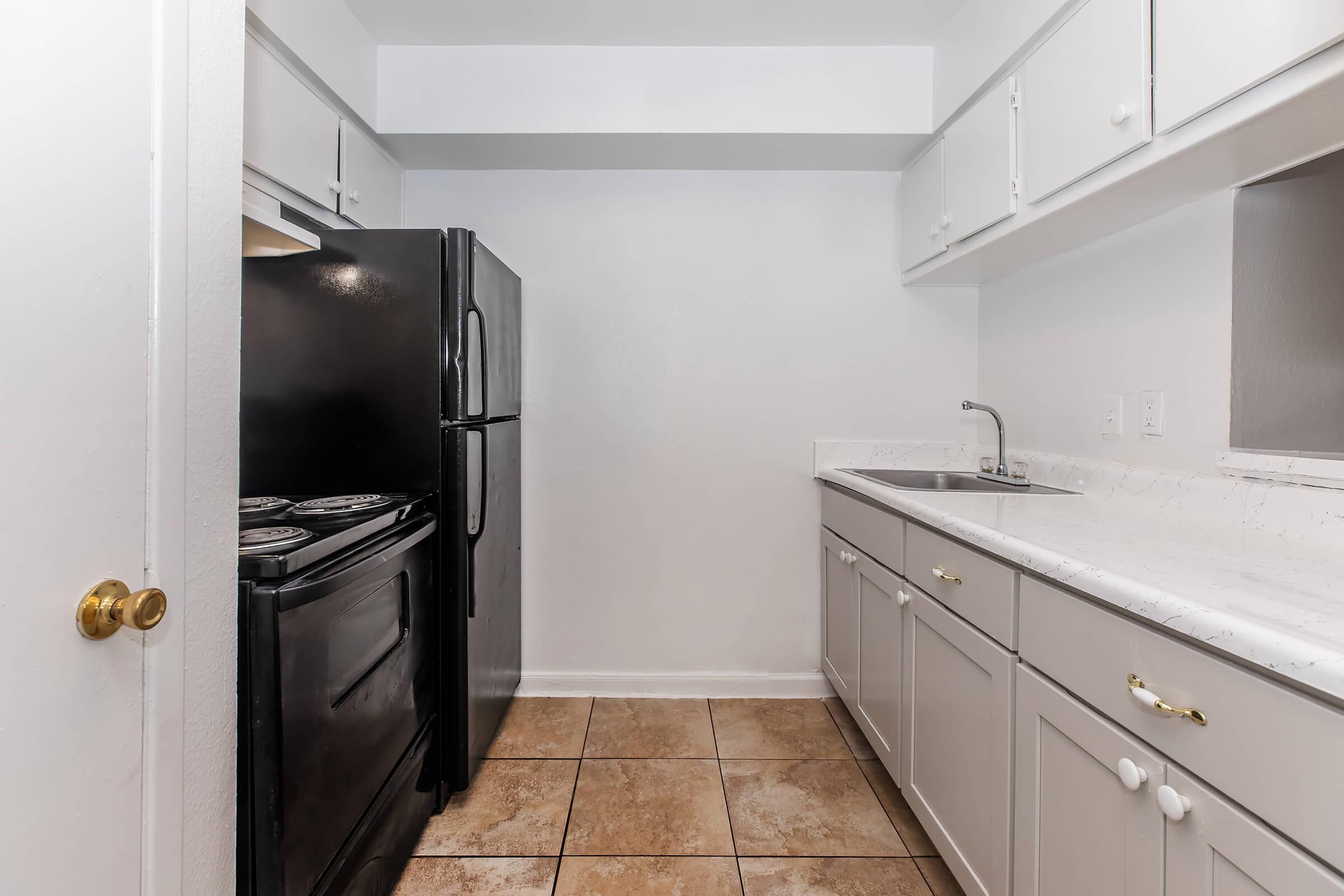 A small kitchen featuring white cabinetry, a black refrigerator, and a black stove. The countertop is light-colored with a sink. The floor is tiled in a beige pattern, and the walls are painted white, creating a clean and modern look.