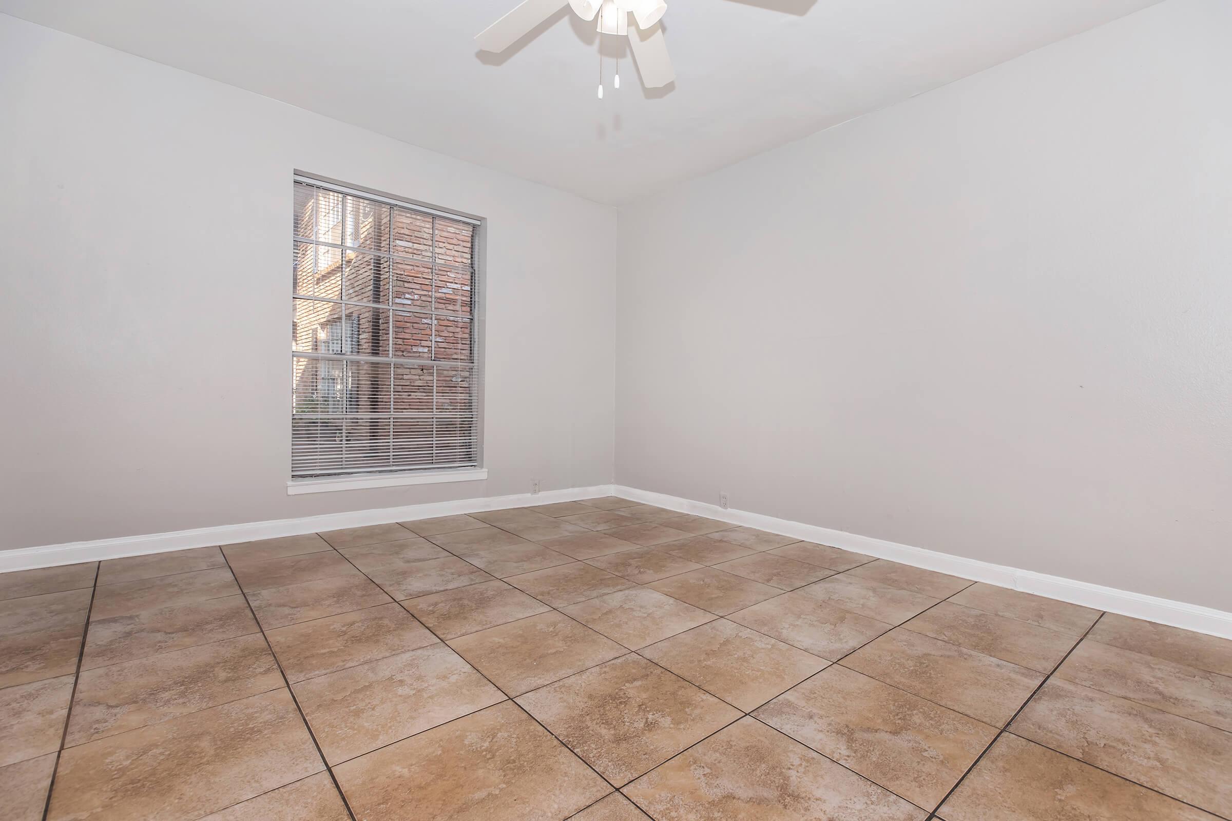 An empty room with light beige tiled flooring and neutral gray walls. A window with blinds is located on the left side, allowing natural light to enter. The ceiling features a circular ceiling fan. The space is clean and airy, suitable for various decorative styles.